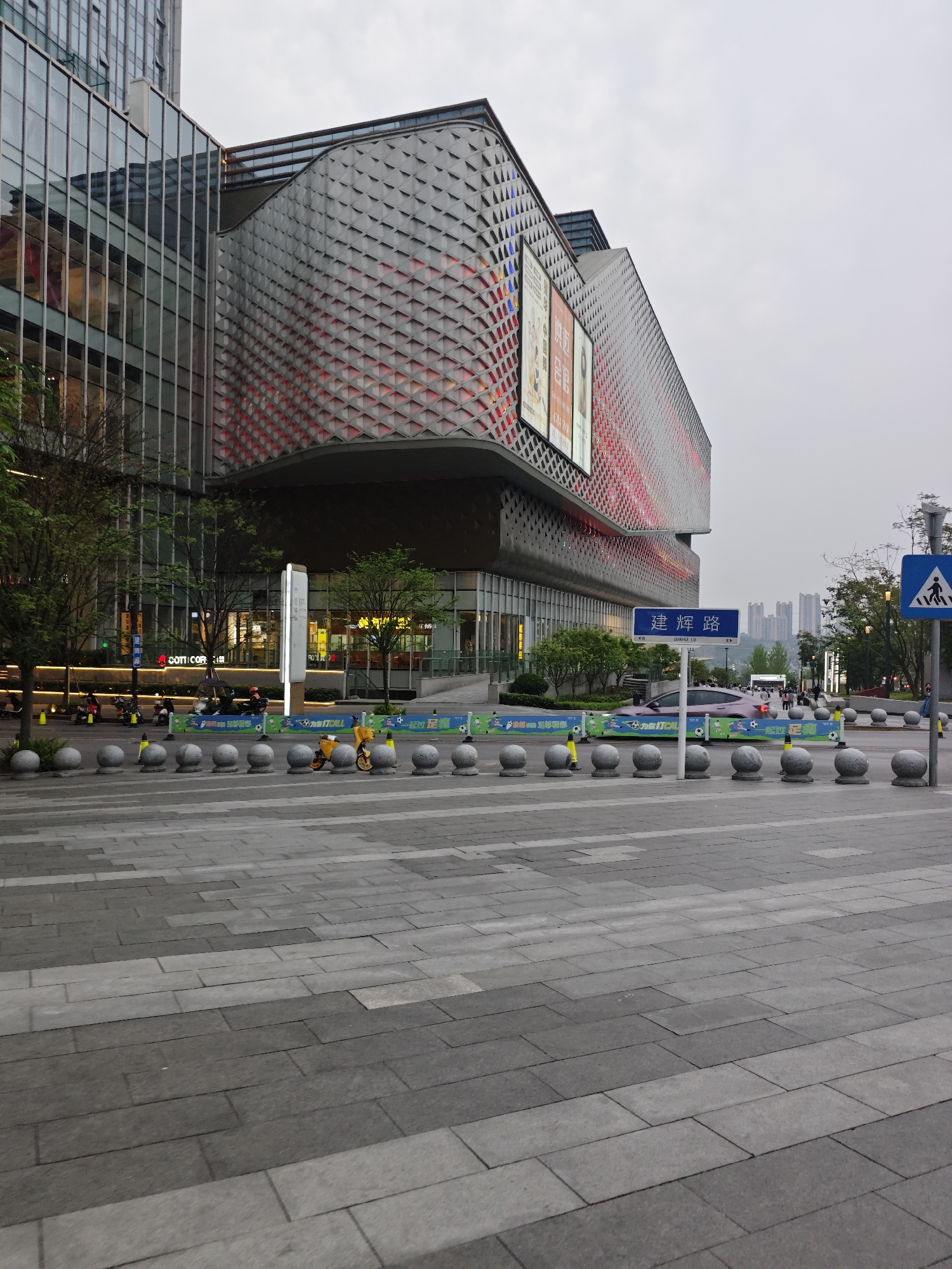 The image depicts an urban scene in Liangjiang, China. A modern building with a tessellated facade dominates the left side of the frame, adjacent to a glass-clad structure. Across a wide, paved pedestrian area, a row of spherical bollards acts as a barrier. A street sign in Chinese is visible to the right, near a pedestrian crossing sign. In the background, a cluster of high-rise buildings rises in the distance. A person on a yellow scooter and a purple car are also in the shot, in the background. The sky is overcast, suggesting a grey day. The paving stones are arranged in a linear pattern, extending from the bottom of the frame toward the building.