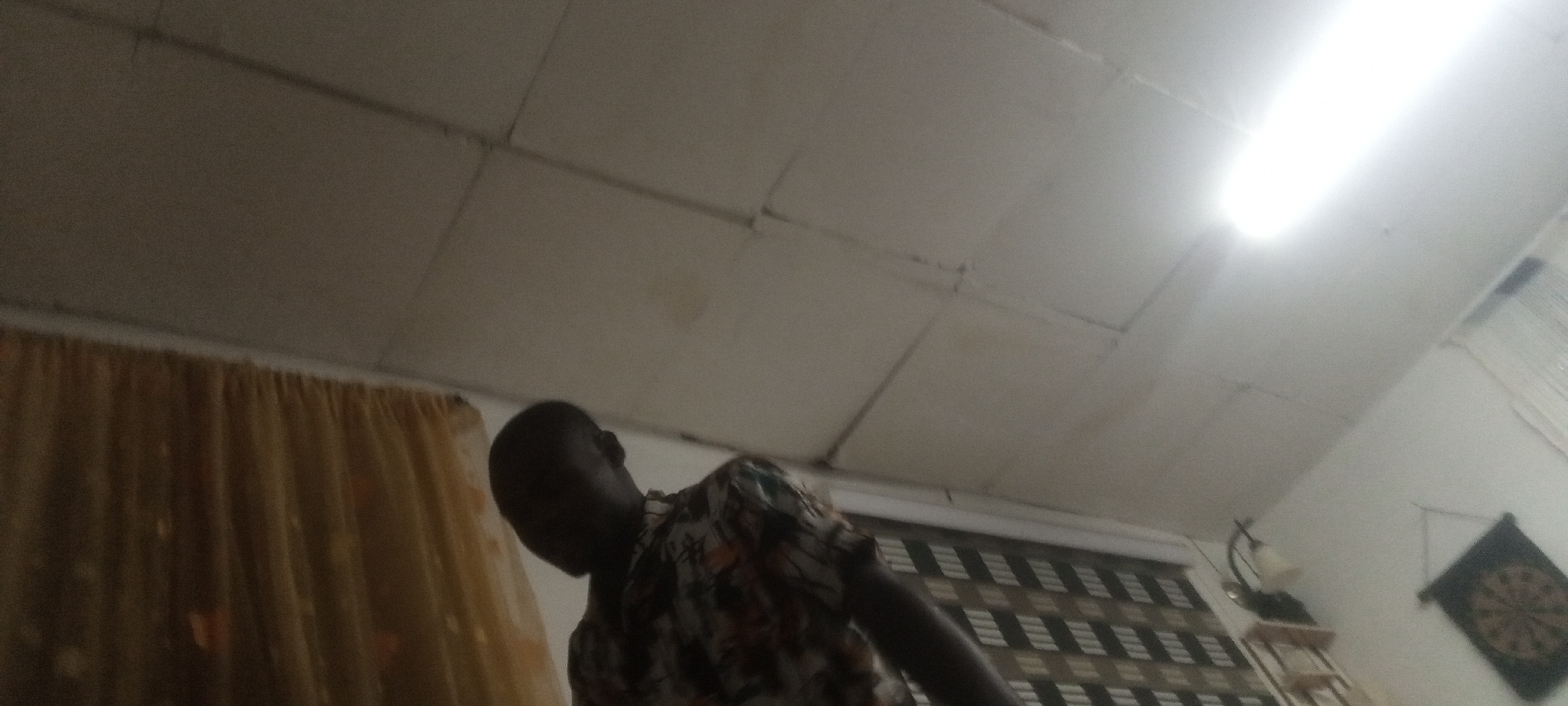 The image shows a room with a fluorescent light fixture illuminating the ceiling. A section of gold-colored curtains is visible on the left. A man wearing a patterned shirt is partially visible in the foreground, his head bowed. In the background, there are horizontally striped blinds covering a window and a dartboard hanging on the wall. The location is Unknown, Unknown.