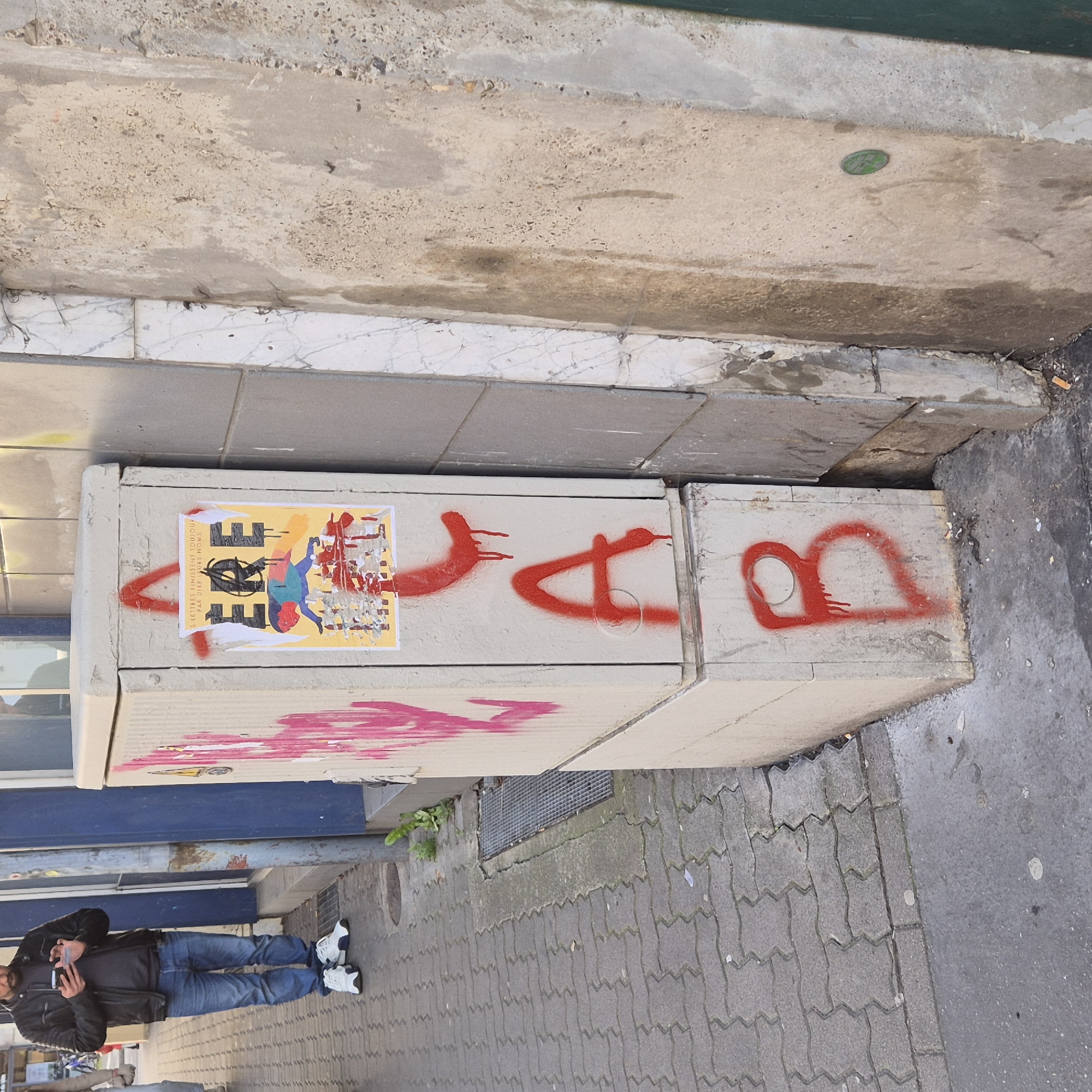 Here is a description of the image:
The image depicts a street scene in Strasbourg, France, with a focus on a set of rectangular concrete blocks serving as a makeshift barrier or seating area on the sidewalk. The blocks are tagged with red graffiti, including what appears to be stylized lettering. A colorful poster is affixed to the side of one of the blocks.
A person is standing on the sidewalk facing the concrete blocks. The person is dressed in jeans and a dark jacket, and is holding a mobile phone in their hand. The sidewalk is paved with a grey interlocking brick pattern. The background includes a curb, a street, and the facade of a building. A grate in the sidewalk is also visible.