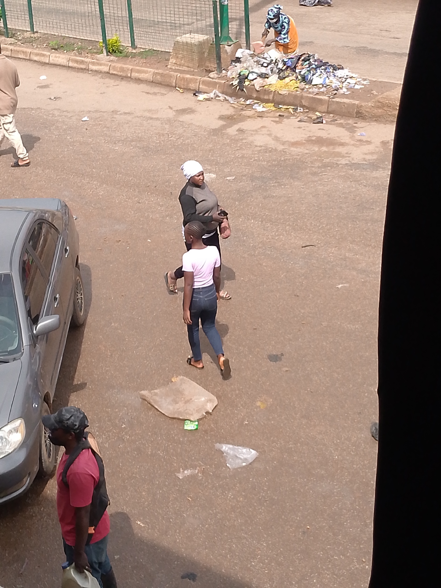 This is a high-angle, medium shot captured in Bukuru, Nigeria, depicting a street scene with several individuals and a car. The foreground is dominated by a grey sedan, parked on the right side of a paved road. To the left of the car, a man wearing a dark camouflage baseball cap, a red t-shirt, and blue jeans is visible, holding a light-colored plastic jerrycan in his left hand and carrying a black backpack. He appears to be walking away from the viewer.
In the middle ground, two women are present. One woman, wearing a white headscarf and a dark long-sleeved top with lighter colored panels, stands with her back to the camera. To her right and slightly in front, a younger woman in a light pink t-shirt and dark blue jeans walks towards the right edge of the frame, wearing sandals.
The background reveals a pile of refuse on the side of the road, with a woman in an orange skirt and patterned headscarf tending to it, holding a pink object, possibly a tool or container. Beyond the refuse, a green metal fence encloses an area with some vegetation. The road surface is asphalt, with scattered litter visible throughout the scene. The overall lighting suggests it is daytime, with shadows cast by the car and people.
