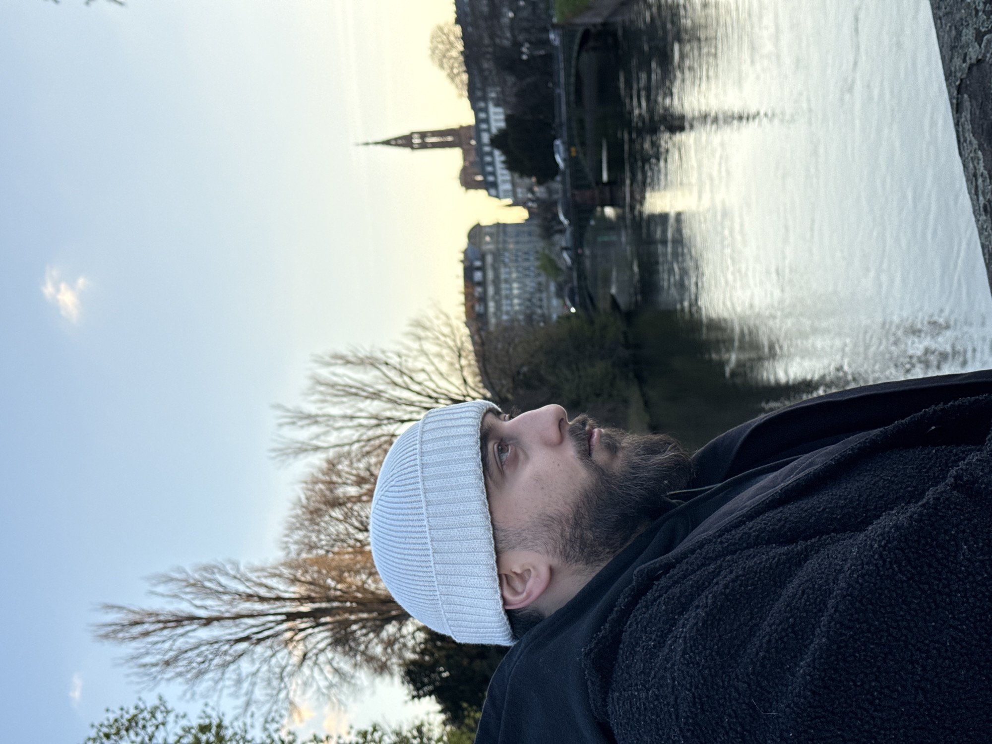 The image depicts a male individual in a profile view, positioned on the right side of the frame, overlooking the Ill River in Strasbourg, France, specifically with the Église Saint-Paul de Strasbourg visible in the background.
The subject is a man in his 20s or 30s with a dark beard and dark hair visible under a light grey, ribbed knit beanie. He wears a dark, possibly black, hooded garment beneath a dark, textured, possibly fleece or sherpa-lined outer jacket. His gaze is directed towards the right, out of the frame.
The background features the Ill River, exhibiting reflections on its calm surface. Beyond the river, at least one arch bridge is visible, carrying vehicular traffic. Further in the distance, typical multi-story European buildings with pitched roofs line the riverbanks. The prominent gothic spire of the Église Saint-Paul de Strasbourg stands tall and slender in the mid-right background, rising above the urban landscape. Bare deciduous trees, indicative of late autumn or winter, are visible on both sides of the river. The sky above is light blue with a faint yellow-orange tint near the horizon, suggesting late afternoon or early evening. A small, diffuse white cloud is present in the upper left sky. In the immediate foreground, on the bottom right, a grey, textured stone surface, possibly a bridge railing or embankment wall, is partially visible.