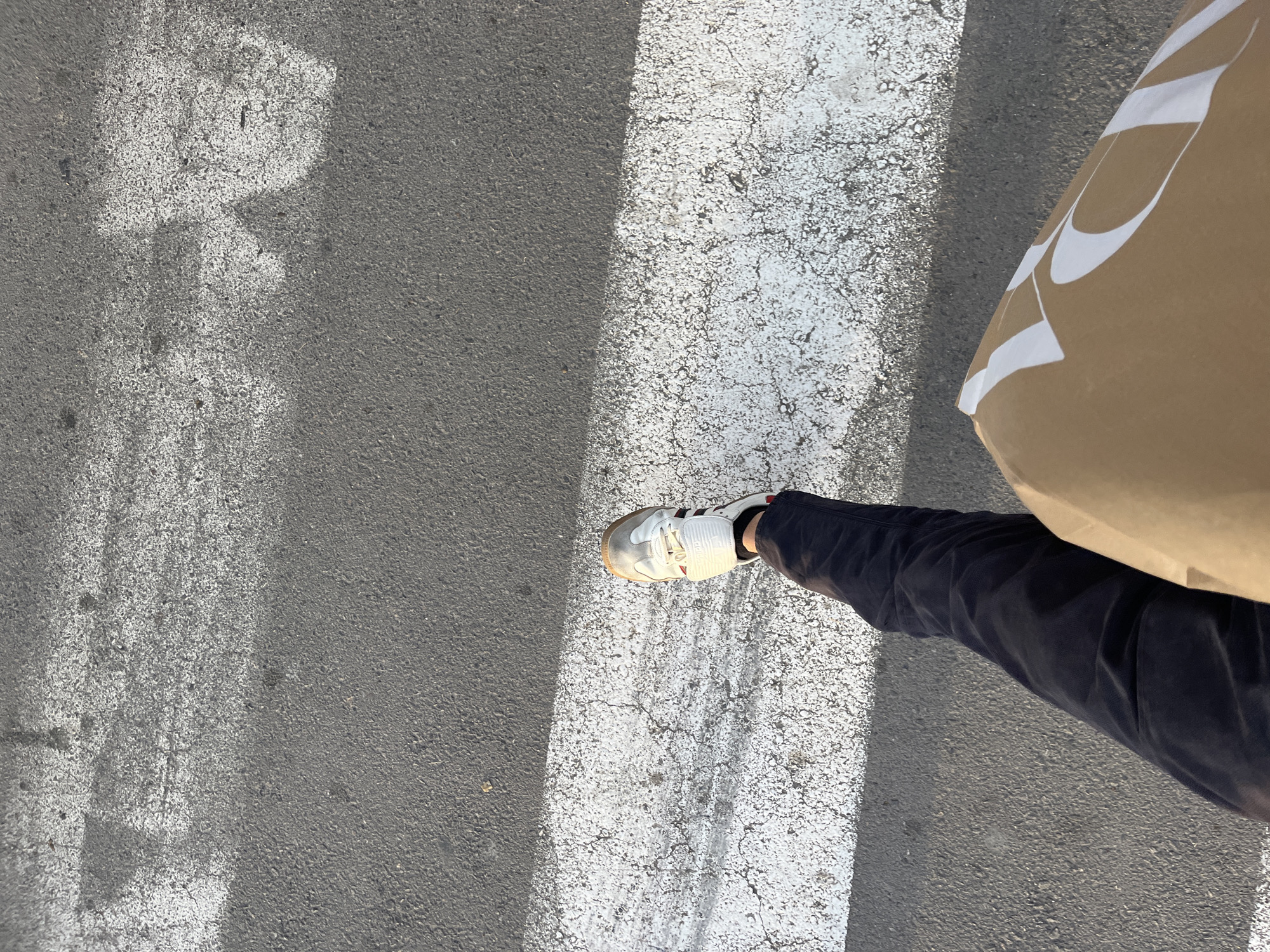 The image provides a high-angle, overhead view of a person's lower leg and foot placed on a textured asphalt road in Torrent, Spain. The setting is outdoors under bright sunlight, evidenced by the distinct, long shadow cast across the ground, dividing the white road marking and the dark asphalt into areas of light and shadow.
The subject is a person, of whom only a leg, foot, and part of an arm (implied by the bag) are visible. They are wearing dark-colored trousers and a pair of white sneakers with subtle dark and red striped accents on the side and a light brown/gum-colored sole. The foot is positioned on a wide, worn white line painted on the road, suggestive of a crosswalk or other traffic marking.
To the right side of the frame, the person is holding a large, light brown paper bag. This bag features prominent, stylized white designs that appear to be abstract letterforms or a brand logo, though specific text cannot be fully deciphered from the visible portions.
The asphalt road surface is dark gray, coarse, and grainy. The white painted lines are visibly aged, showing cracks and some wear. Another faded, irregular white marking is partially visible on the darker asphalt to the left. The overall impression is a snapshot of someone walking or pausing on a sunny day in an urban or suburban environment.