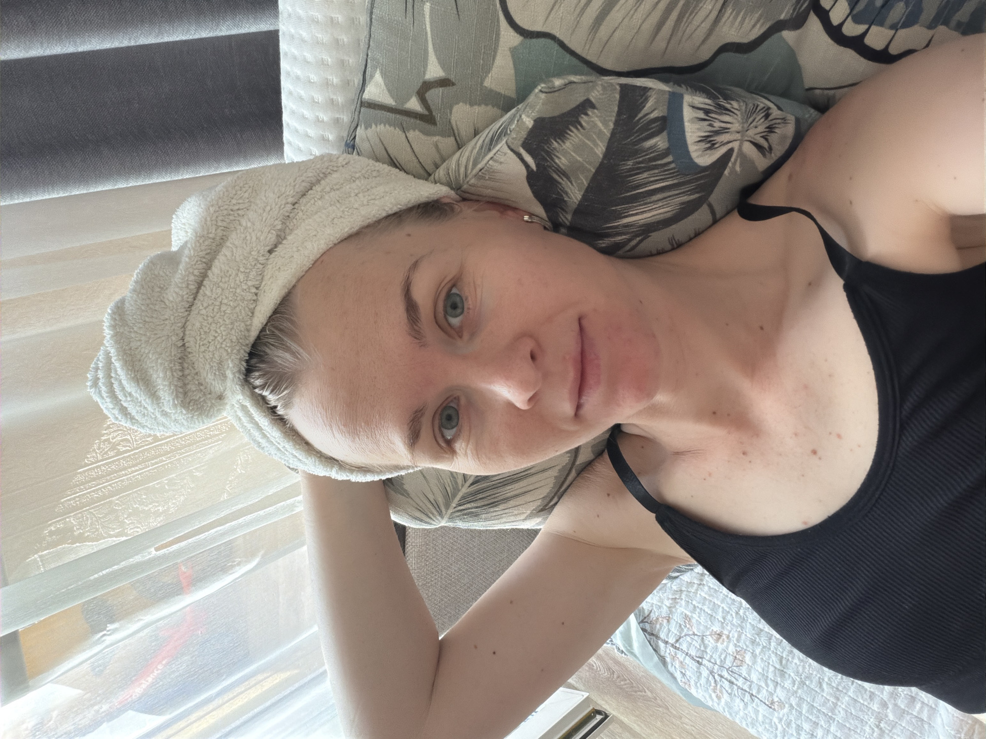 A woman with a towel wrapped around her head is lying on a bed, looking at the camera. She is wearing a black tank top. Her left arm is raised and bent behind her head. She has fair skin and light blue eyes. The background includes a sheer curtain and a patterned pillow. The scene appears to be indoors, likely in a residential setting, such as an apartment or house. Based on the provided information, the location is Khabarovsk, Russia.