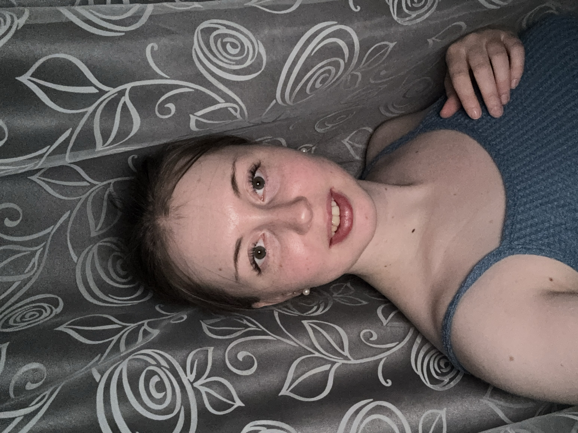 A young woman is depicted lying on a patterned surface, looking directly at the camera with a soft smile. Her head is angled, suggesting she is lying on her left side. She has fair skin, brown eyes, and her brown hair is pulled back. She is wearing a light blue, ribbed top with thin straps. A pearl earring is visible on her left ear. The background consists of a dark grey or black fabric adorned with a prominent white floral and swirling pattern, resembling upholstery or a curtain. Her right hand is visible near the upper right corner of the frame, resting on her shoulder or upper arm. The lighting is soft and even. There is no visible text in the image.