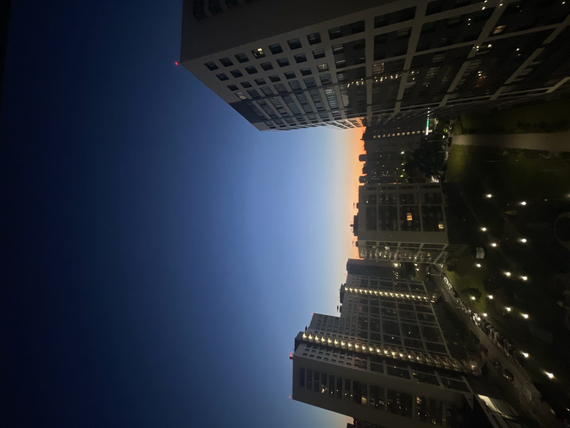 The image captures a twilight view of a residential area in Sinyavino, Russia. Several tall, modern apartment buildings dominate the scene, with windows illuminated from within, indicating evening occupancy. The sky transitions from a deep blue at the top to a warm orange and yellow near the horizon, typical of sunset or sunrise. In the foreground, a dark, paved area with scattered streetlights suggests a pathway or courtyard. Cars are parked along a street adjacent to the buildings. The overall environment is urban, with a focus on residential architecture. There are no visible people or animals in the frame.