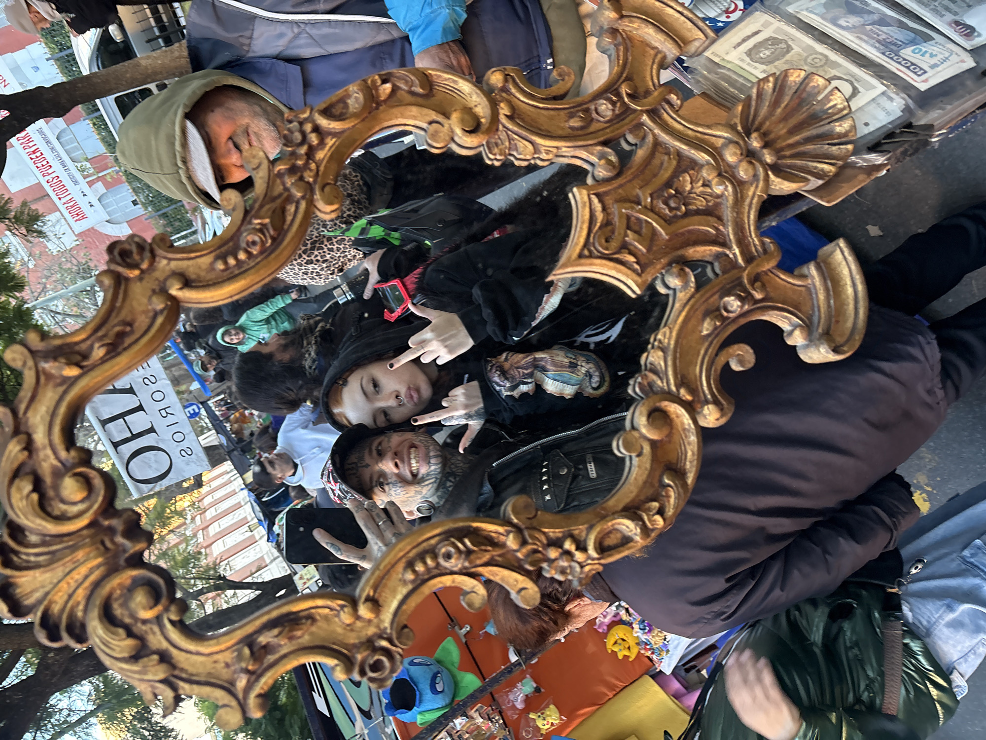 The image shows a street scene captured through an ornate, gold-colored mirror frame. In the reflection, two individuals are visible: one with extensive facial tattoos and the other with facial piercings, both gesturing with their hands. The mirror is positioned amongst various items for sale on a blanket-covered surface, including plush toys, stickers, and foreign currency. Other individuals, including one wearing a hooded jacket, can be seen in the background, along with a building with a sign that reads "AORA TODOS PUEDEN PAR." The location is Buenos Aires, Argentina.