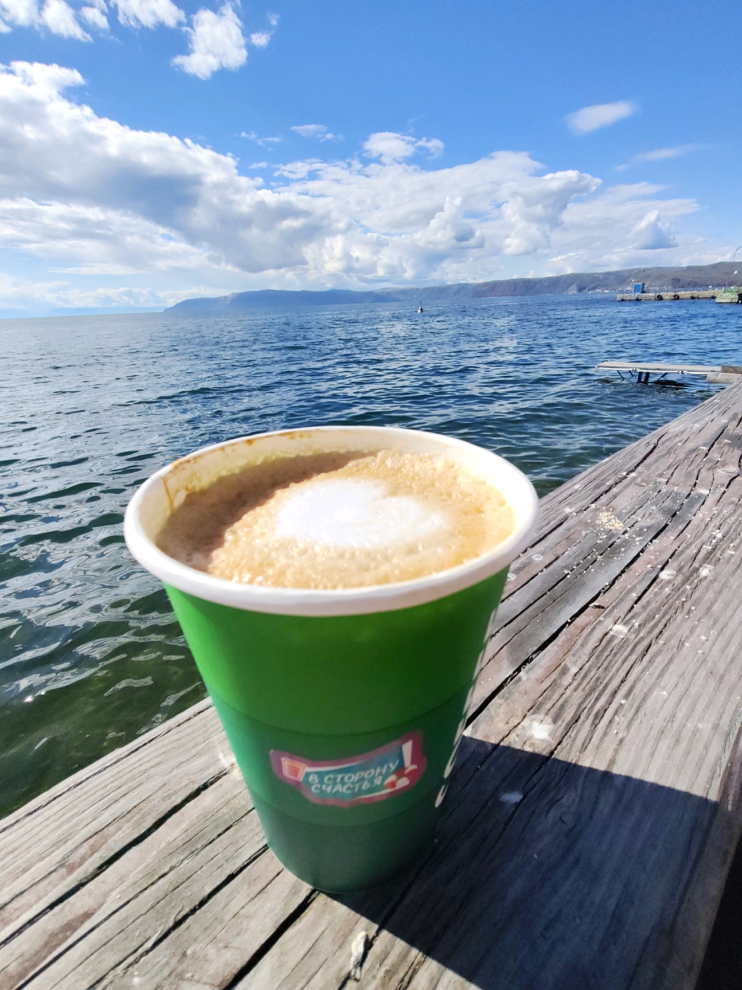 A close-up shot showcases a bright green paper cup filled with coffee, resting on a weathered wooden pier. The coffee has a foamy, creamy topping. The pier is positioned along the edge of a vast body of water, likely Lake Baikal, given the mention of Listvyanka, Russia. The water is a deep blue, with ripples reflecting the bright blue sky, which is dotted with fluffy white clouds. In the distance, a hilly shoreline is visible, and a small dock extends into the water on the right side of the frame. A small pink and red sticker with Russian text is affixed to the coffee cup. The overall scene evokes a sense of relaxed enjoyment in a natural setting.