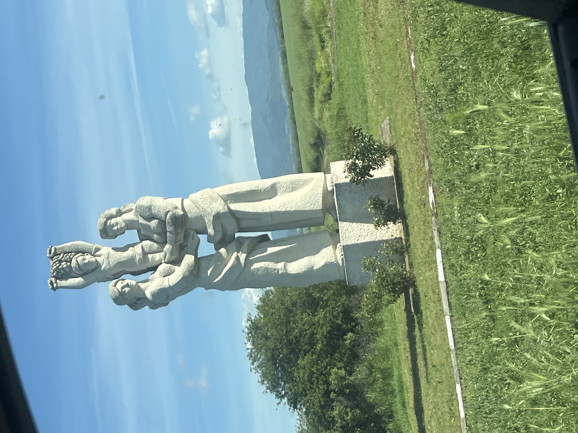 The image shows a stone monument, likely of socialist realism style, depicting three figures stacked atop one another. The monument is situated alongside a road or path in a rural or semi-rural location, possibly the city of Brezovo, Bulgaria, based on the user's query. In the background are fields of tall green grass and in the distance, some mountains. The sky is blue with a few scattered clouds. The photo appears to have been taken from inside a vehicle.