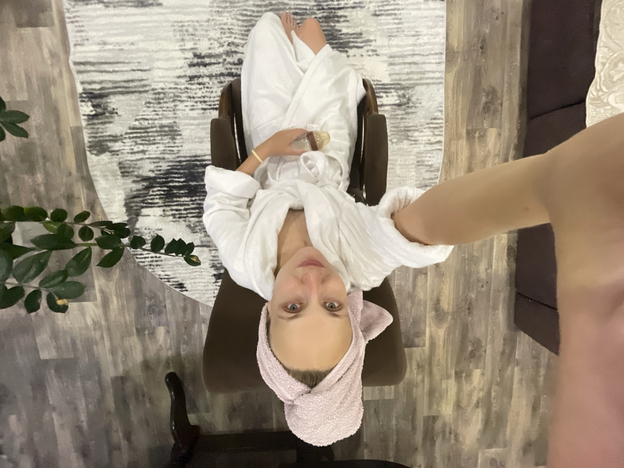 A person sits in a brown armchair, wearing a white bathrobe and a pink towel wrapped around their head. They are holding a baby bottle in their right hand and looking at the camera. The person's arm is extended, suggesting they are taking a selfie. The background shows a wooden floor, a patterned rug, and a plant. The scene appears to be indoors, possibly in a home or hotel room. The city of Sharkan, Russia is not depicted or suggested in the image.