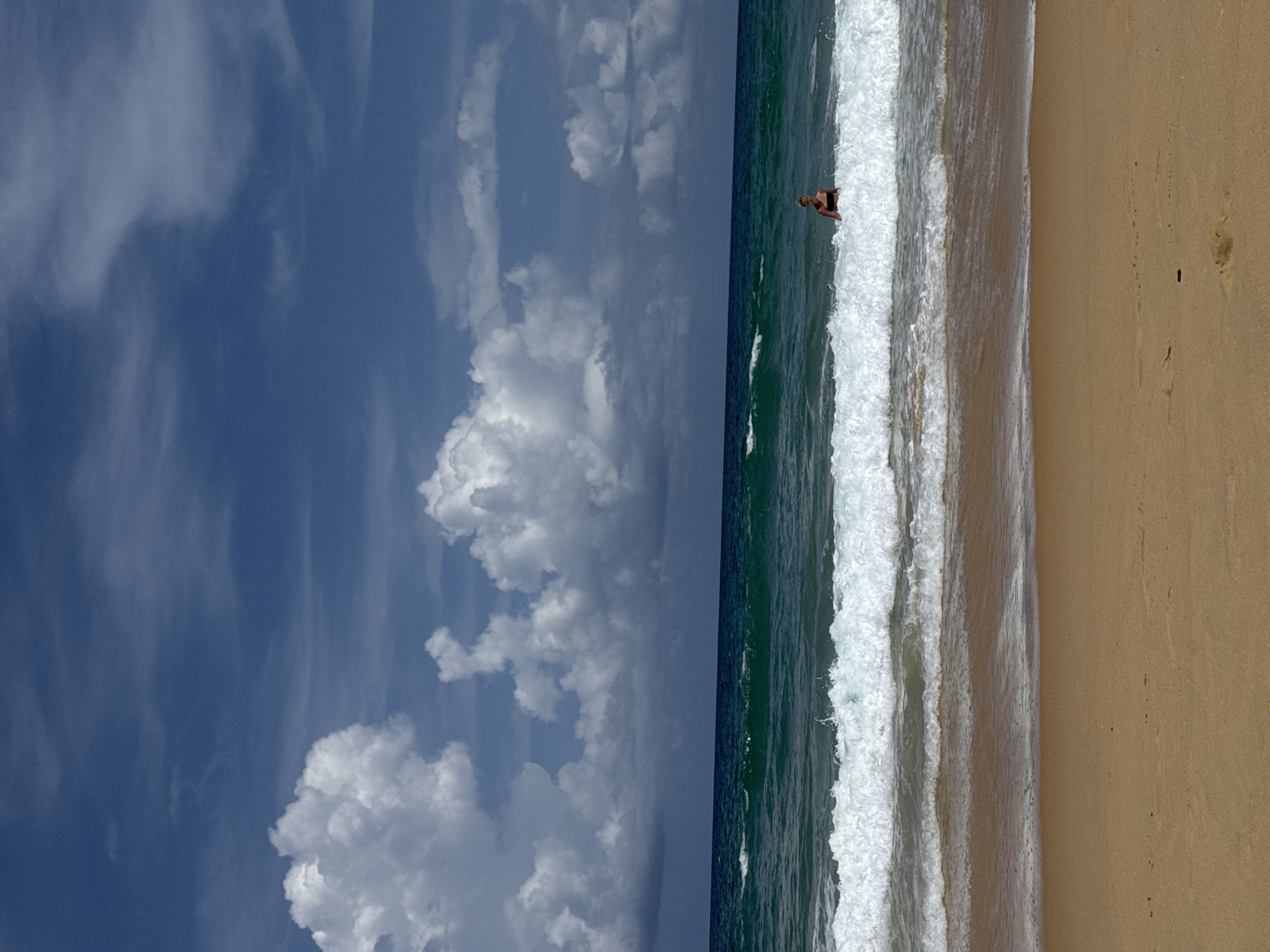 A woman in a black bikini stands in the ocean water, partially submerged, with foamy waves breaking around her. She is facing towards the viewer, with a calm expression. The scene is set on a sandy beach, with the ocean stretching out to the horizon. The sky is a deep blue, with fluffy white clouds scattered across it. The water is a vibrant turquoise, with white caps on the waves. The beach is golden sand, with a few small footprints visible. The location is Karon Beach, Karon, Thailand.