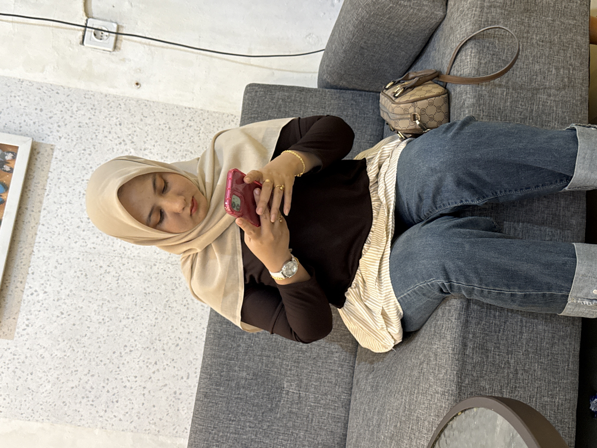 A woman wearing a beige hijab, dark brown long-sleeved shirt, and blue jeans is sitting on a gray sofa while looking at a pink mobile phone. She is wearing a gold bracelet, rings on her fingers, and a silver wristwatch. A small brown purse is resting on the sofa beside her. The scene is indoors with a white speckled wall visible in the background, with an electrical outlet. The exact location within the city of Lhokseumawe, Indonesia cannot be determined from the image.