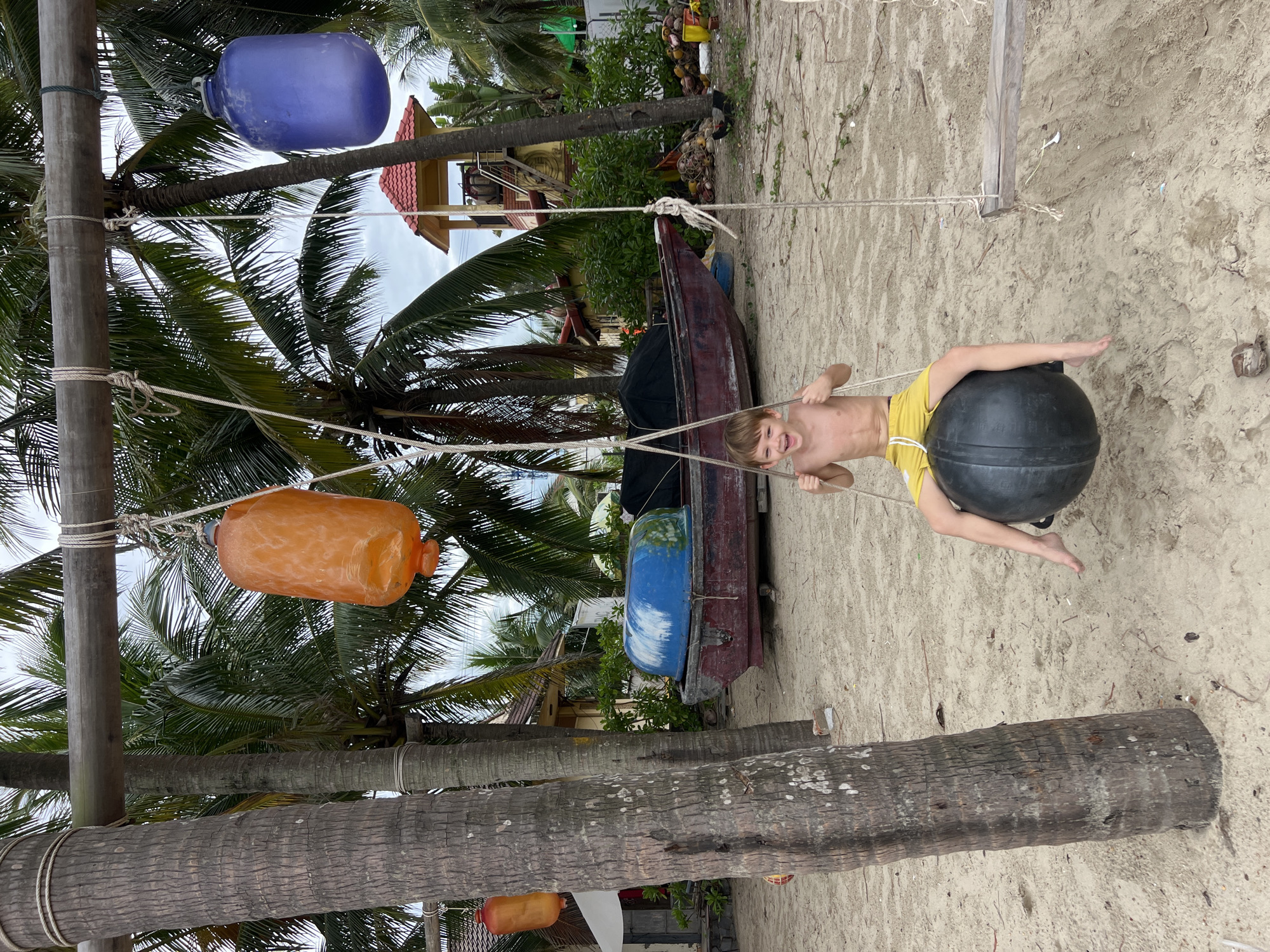 A young boy sits on a large black sphere suspended by ropes, as if on a swing, on a sandy beach. He is shirtless and wearing yellow shorts, with a wide, excited smile on his face. The swing is attached to a wooden beam supported by two palm trees. Orange and blue buoys hang from the same beam. In the background, a traditional Vietnamese fishing boat is pulled ashore, and a yellow building with a red roof stands among lush greenery. This scene is likely located on a beach in Hoi An, Vietnam, a city known for its beaches, ancient town, and vibrant maritime culture. The overall atmosphere is one of youthful joy and relaxation in a tropical setting.