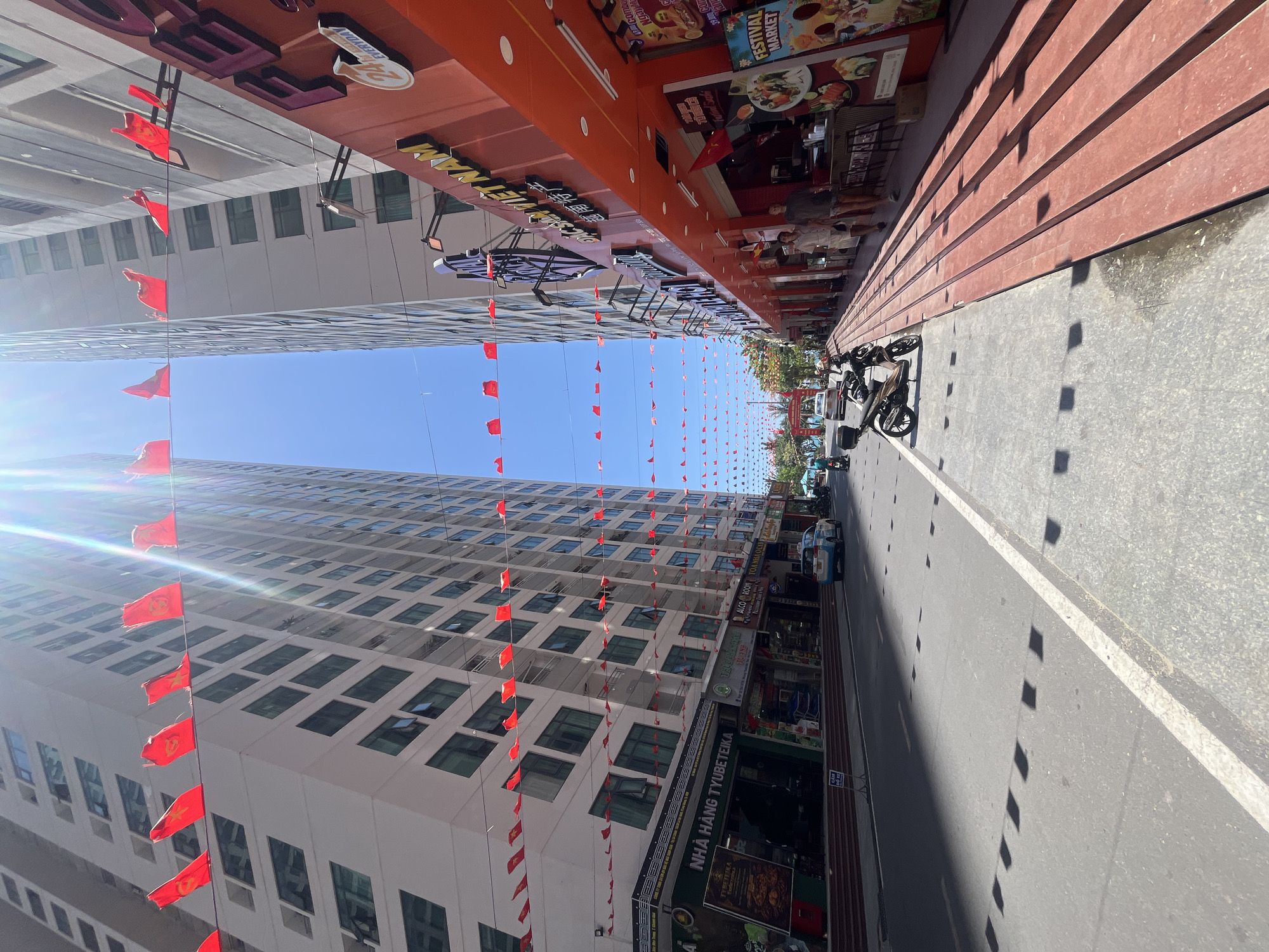 This street-level photograph captures a street scene in Bac Nha Trang Ward, Vietnam. The perspective is set along the street, looking towards the horizon.
On the left, a tall building with a grid-like pattern of windows ascends into the clear blue sky, with numerous red flags hung along its facade. The flags extend across the sky, linking to the buildings on the right side of the street.
On the right side of the street are commercial establishments, including restaurants and shops, with signboards displaying Vietnamese and other languages. Among them are: NHÀ HÀNG TYUBETEIKA, AEGERS, ALCO ROOM TOA NHAOCI, TYBETEIKA, FESTIVAL MARKET. Motorbikes are parked along the sidewalk. The street pavement is gray, with visible lane markings and shadows.