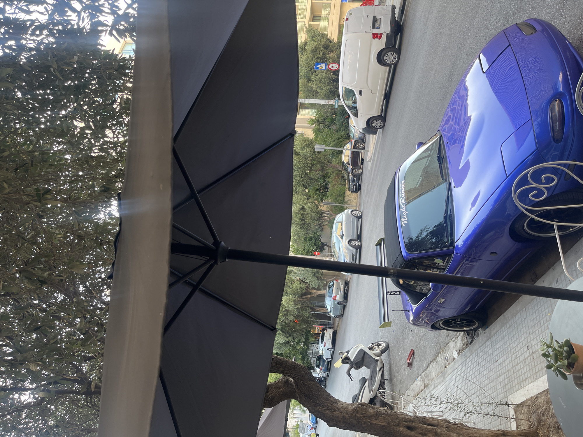 The image shows a street scene in Athens, Greece, viewed from beneath a large black umbrella. In the foreground, a vibrant blue convertible car, possibly a Mazda Miata with a prominent rear spoiler, is parked on the right side of the street. The car has a black soft top and a sticker on the windshield that reads "Night Runner". To the left of the car, a white scooter is parked on the sidewalk next to a tree with a textured trunk. The sidewalk appears to be paved with square tiles.
Across the street, several other vehicles are parked, including a white van, a dark SUV, and various sedans in different colors. The street is lined with trees and buildings, suggesting an urban environment. The lighting indicates it is daytime, with sunlight filtering through the leaves of the trees, creating dappled shadows. A small potted plant sits on a table in the lower left corner of the frame, partially obscuring the view of the sidewalk.