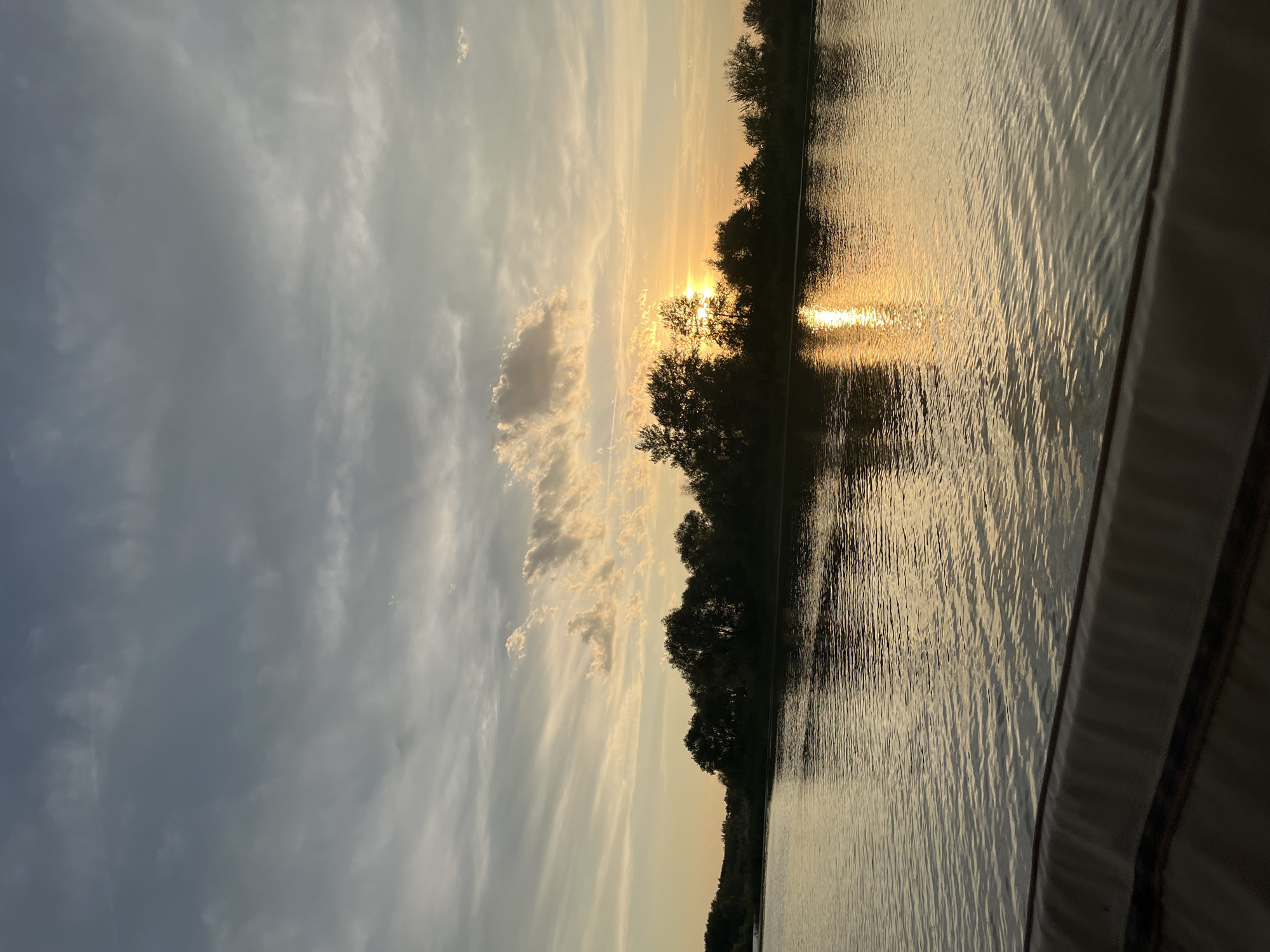 The image depicts a sunset over a body of water, likely a river or lake, with a silhouetted treeline in the background. The sky is filled with clouds of varying shades of gray and white, with patches of orange and yellow light from the setting sun breaking through. The sun itself is partially obscured by trees, casting a warm, golden reflection on the rippling water. In the foreground, a dark, textured surface, possibly the edge of a boat or pier, is visible. The scene is peaceful and natural, evoking the quiet beauty of dusk. The specific location within Шумейка, Russia, is not discernible from the visual information provided.