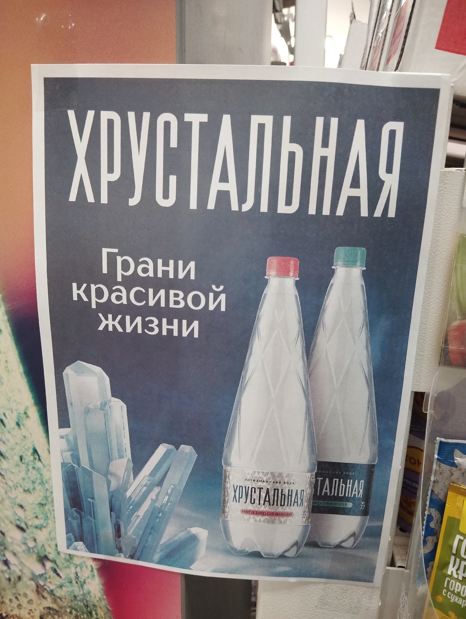 A poster for ХРУСТАЛЬНАЯ water, likely in a store in Krasnogorskoye, Russia. The poster has a gray background with white text reading "ХРУСТАЛЬНАЯ" at the top and "Грани красивой жизни" below. Two bottles of the water are displayed, one with a red cap and labeled "НЕГАЗИРОВАННАЯ" (non-carbonated), and the other with a teal cap and labeled "ГАЗИРОВАННАЯ" (carbonated). The bottom left corner of the poster features an illustration of crystals. Parts of a cereal box and other items are visible to the right.
