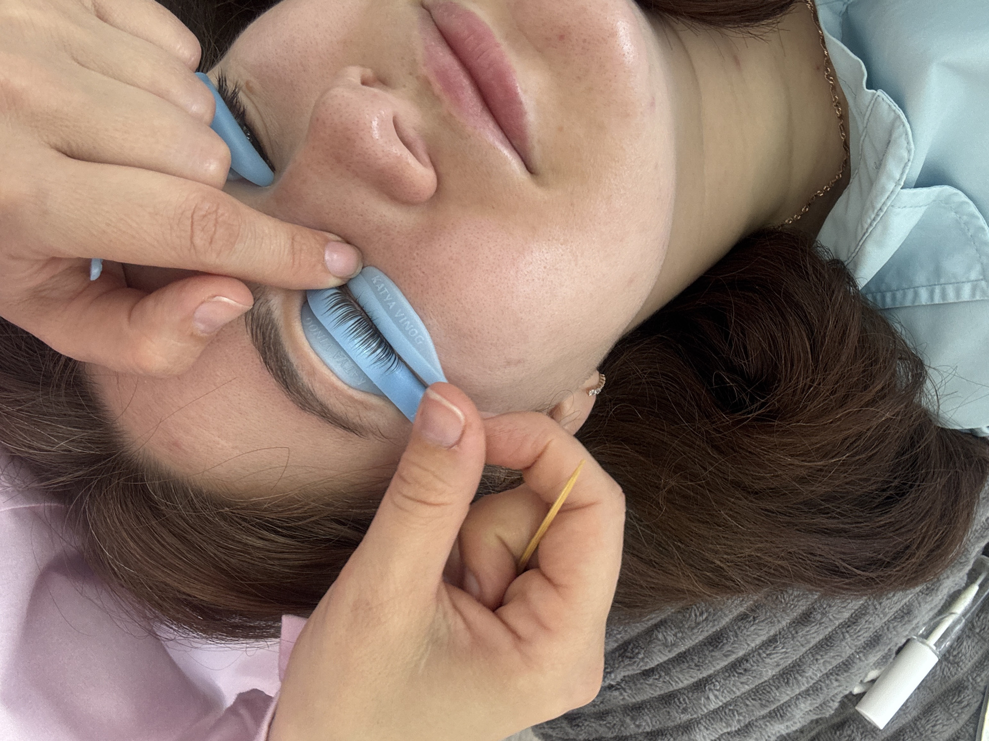 The image depicts a close-up, high-angle shot of a young woman's face as she undergoes a lash lift procedure. Her eyes are closed, and her lashes are being lifted and separated using blue silicone shields and what appears to be a wooden toothpick. Two sets of hands, presumably belonging to a lash technician, are actively working on her lashes. One hand holds the toothpick, and the other manipulates the silicone shield. The woman has light skin with freckles and is wearing a light blue collared shirt and a delicate gold chain necklace. The background is blurred, showing a pink fabric, a grey textured blanket, and what appears to be a white tube of product, possibly related to the beauty treatment. The environment suggests an indoor setting, likely a beauty salon. There is no discernible information in the image to associate this scene with the city of Chaikovsky, Russia.