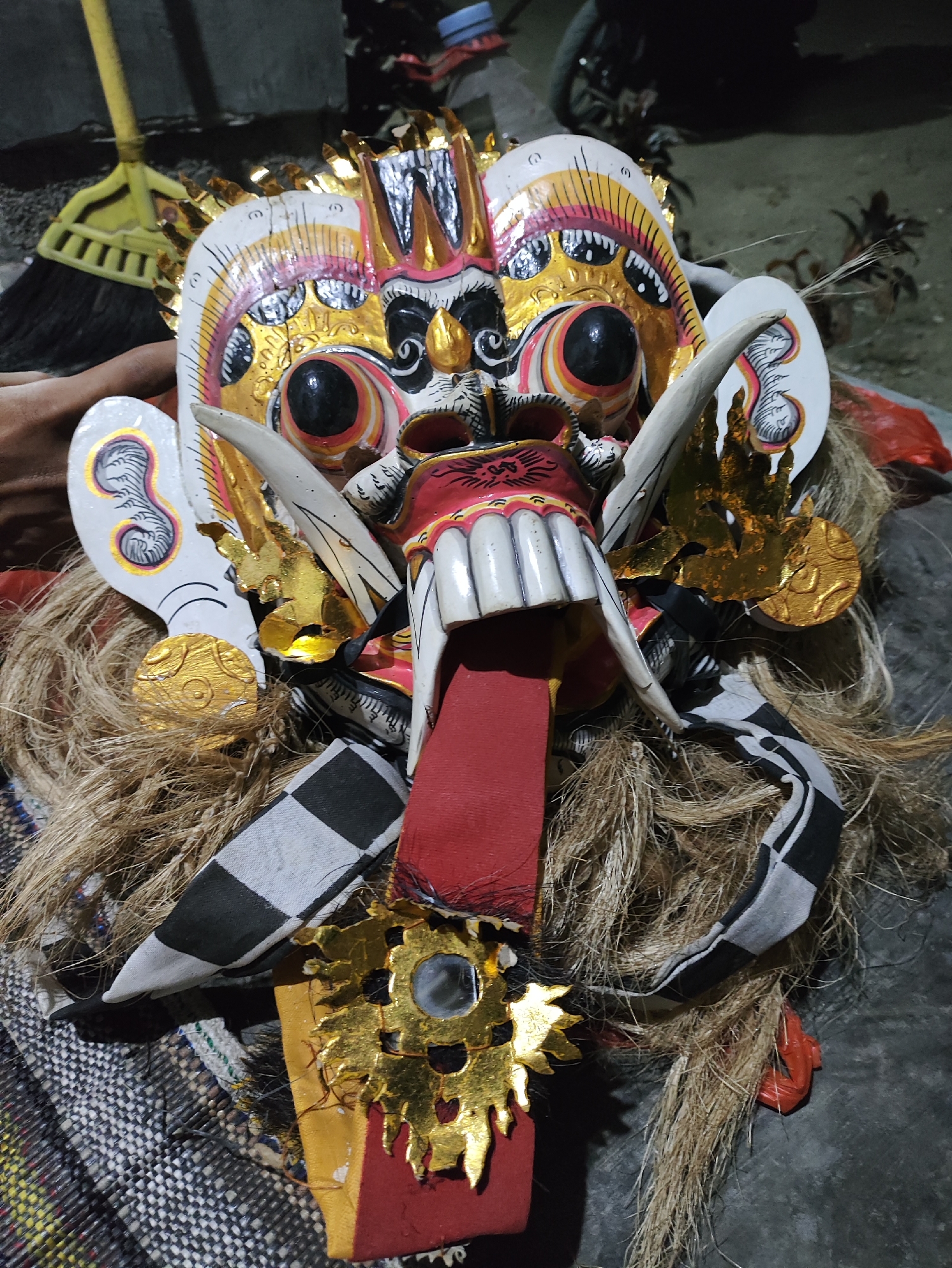 This is a close-up, vertical, full-shot photograph of a colorful, ornate mask, likely a Barong mask from Bali, Indonesia, which is known for its rich cultural heritage and traditions. The mask is detailed with bright red, white, yellow, and black paint, and adorned with gold leaf accents. It features prominent, bulging eyes with black pupils, sharp, white teeth, and a long red tongue protruding from its mouth. The mask also has a thick mane of coarse, light brown fibers cascading around it, and is decorated with gold embellishments, including circular discs and flame-like shapes. A checkered black and white fabric band wraps around the base of the mask, and a red fabric piece is visible at the bottom. A hand, with light skin tone, is visible on the left side of the image, holding the mask. In the background, to the upper left, a yellow broom with a black bristle head is leaning against a textured surface. The environment appears to be outdoors, possibly in a courtyard or open area, with a dark, rough surface beneath the mask. The lighting suggests it is nighttime or a dimly lit area, casting shadows and highlighting the mask's features. While the image does not explicitly show the city of Kepung, Indonesia, it is consistent with the cultural artifacts and artistic styles found in that region, known for its traditional crafts and performances.