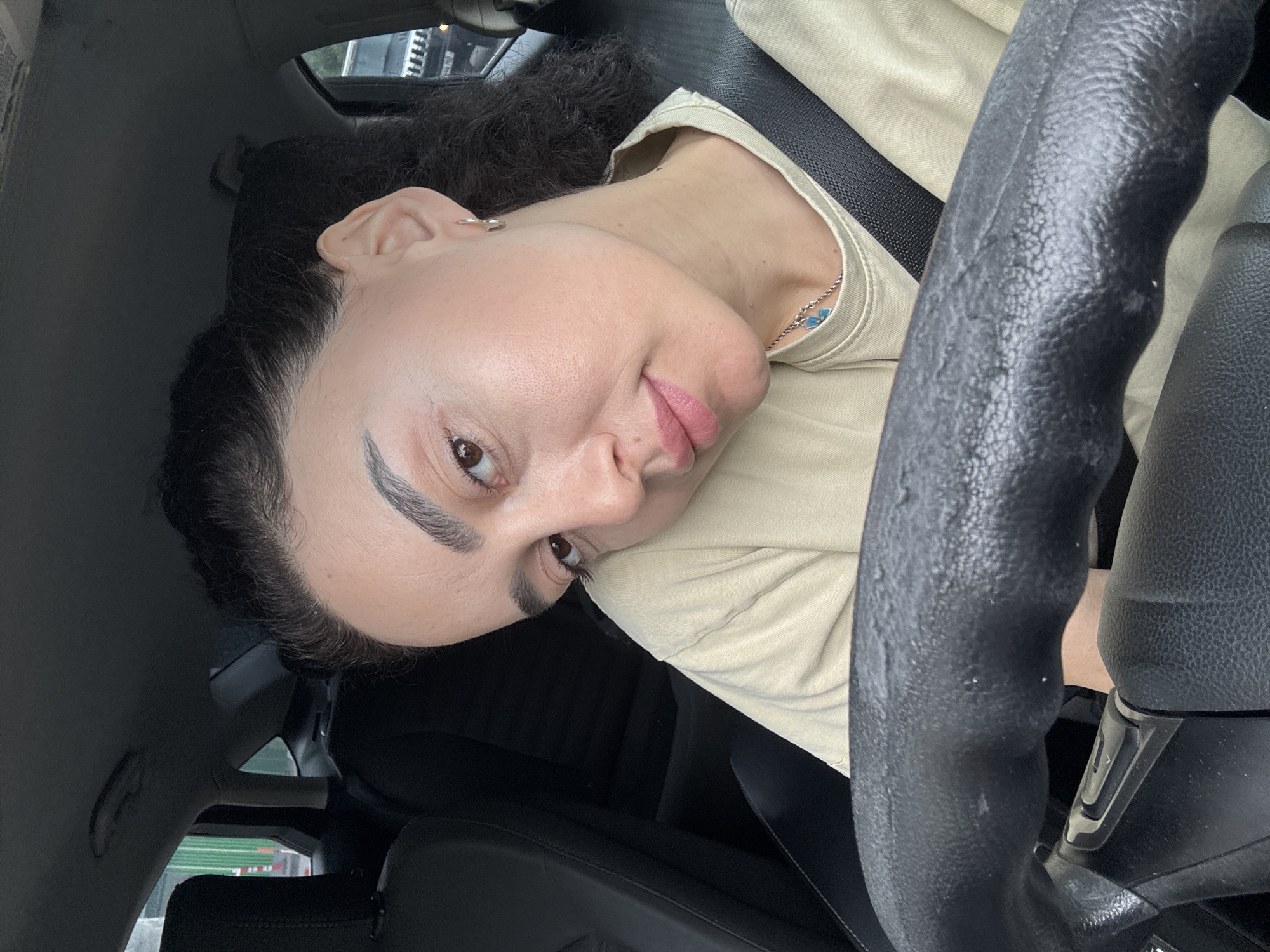 A woman is positioned in the driver's seat of a car. She is wearing a light-colored shirt and a seatbelt. Her hair is dark and curly. She has a necklace with blue stones. The car has a black steering wheel and black leather seats. The location might be Tbilisi, Georgia, but there is insufficient information to confirm this with certainty.
