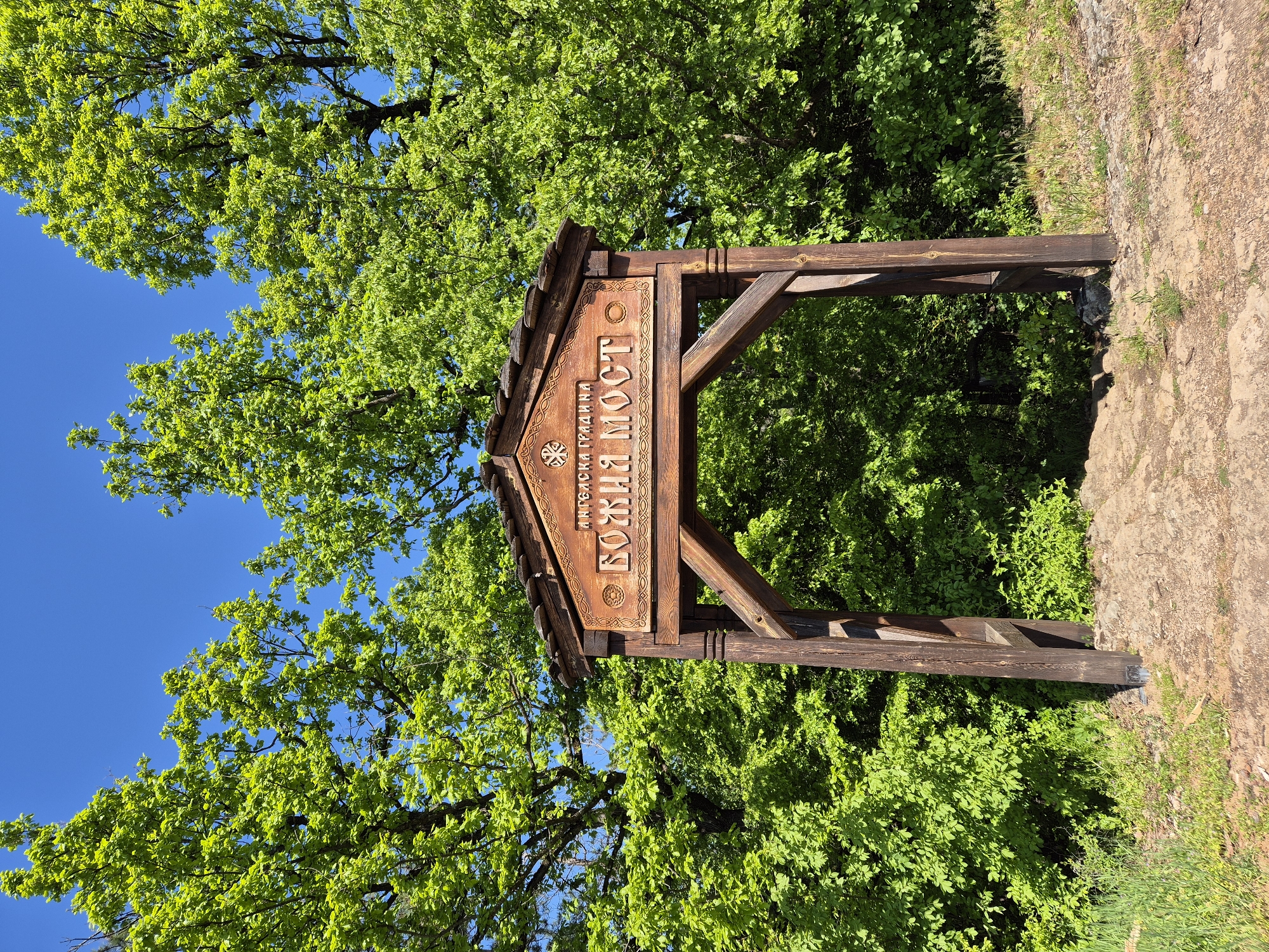 The image shows a wooden archway-style sign in a natural setting, likely a park or forest, based on the abundant greenery. The sign displays text in Cyrillic script, specifically reading "Божия Мост" (Bozhia Most), which means "God's Bridge," alongside the text "МИРБАСКА ТРАДИЦИЯ." The environment consists of lush green foliage, possibly trees or bushes, with a patch of earth or dirt visible on the right side. The sky is a clear blue, visible behind the foliage on the left. The sign appears to be located in or near Lilyache, Bulgaria.