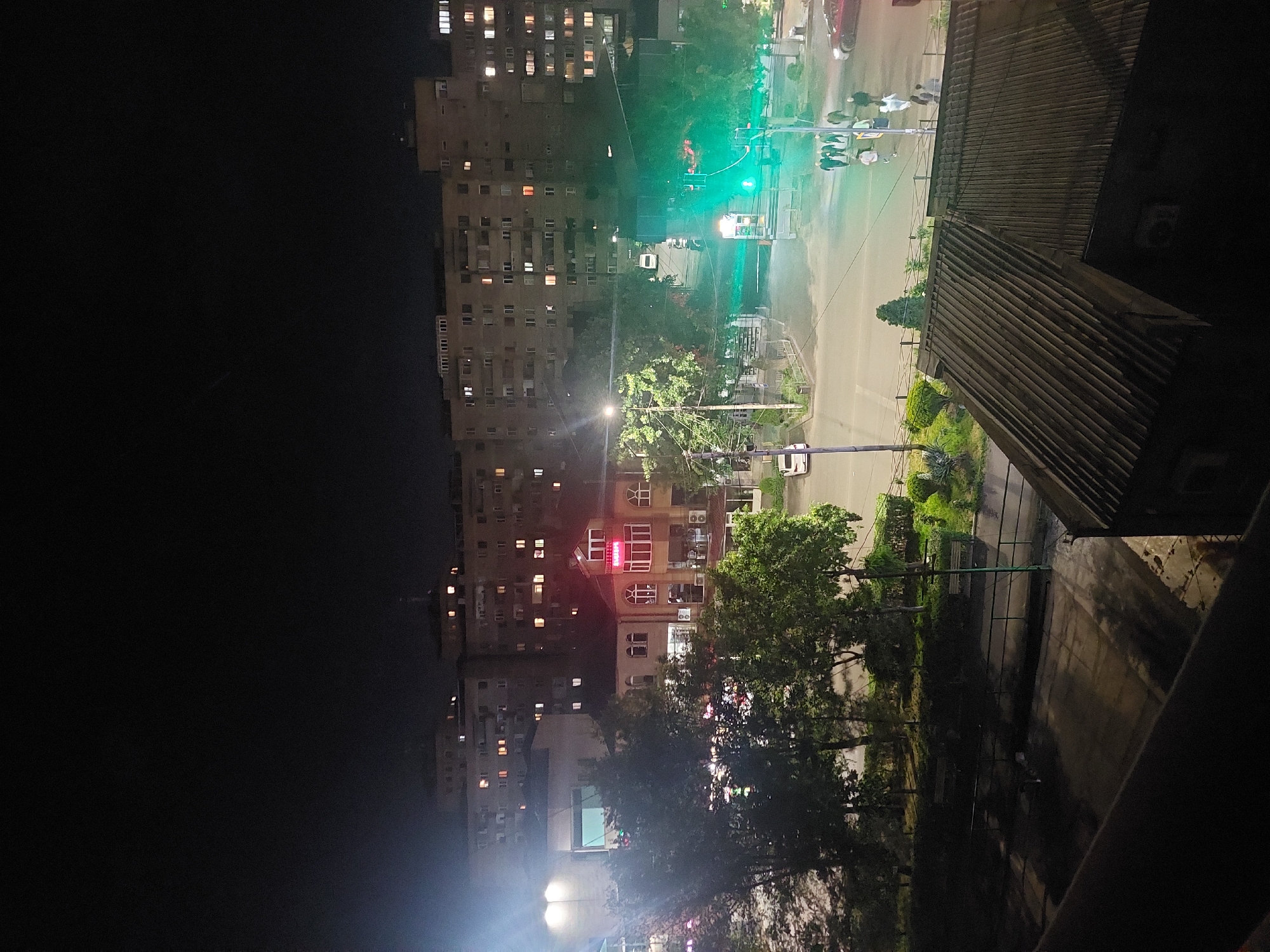 The image captures a vibrant nocturnal urban scene in Yerevan, Armenia, from an elevated viewpoint. Large, multi-story residential buildings with numerous illuminated windows line the left side of the frame, indicating active living spaces. A well-lit street cuts across the middle ground, featuring streetlights and a prominent green traffic signal, suggesting ongoing traffic flow. Several blurry figures, likely pedestrians, are visible near the traffic light, and a white car is partially seen on the road, with a red vehicle further in the distance.
Abundant greenery, including dense trees and bushes, is interspersed throughout the scene, particularly along the street and between buildings, adding natural elements to the urban landscape. On the right side of the street stands a distinctive building with white-framed windows and a prominent red illuminated sign that clearly displays the word "BEAUTY" in white capital letters. In the immediate foreground on the right, a dark, corrugated metal roof and a partial view of a balcony railing indicate the vantage point of the photographer. The time of day is clearly night, with various artificial light sources creating a lively yet tranquil atmosphere. The weather appears clear. Utility poles and wires are visible, crossing the sky.