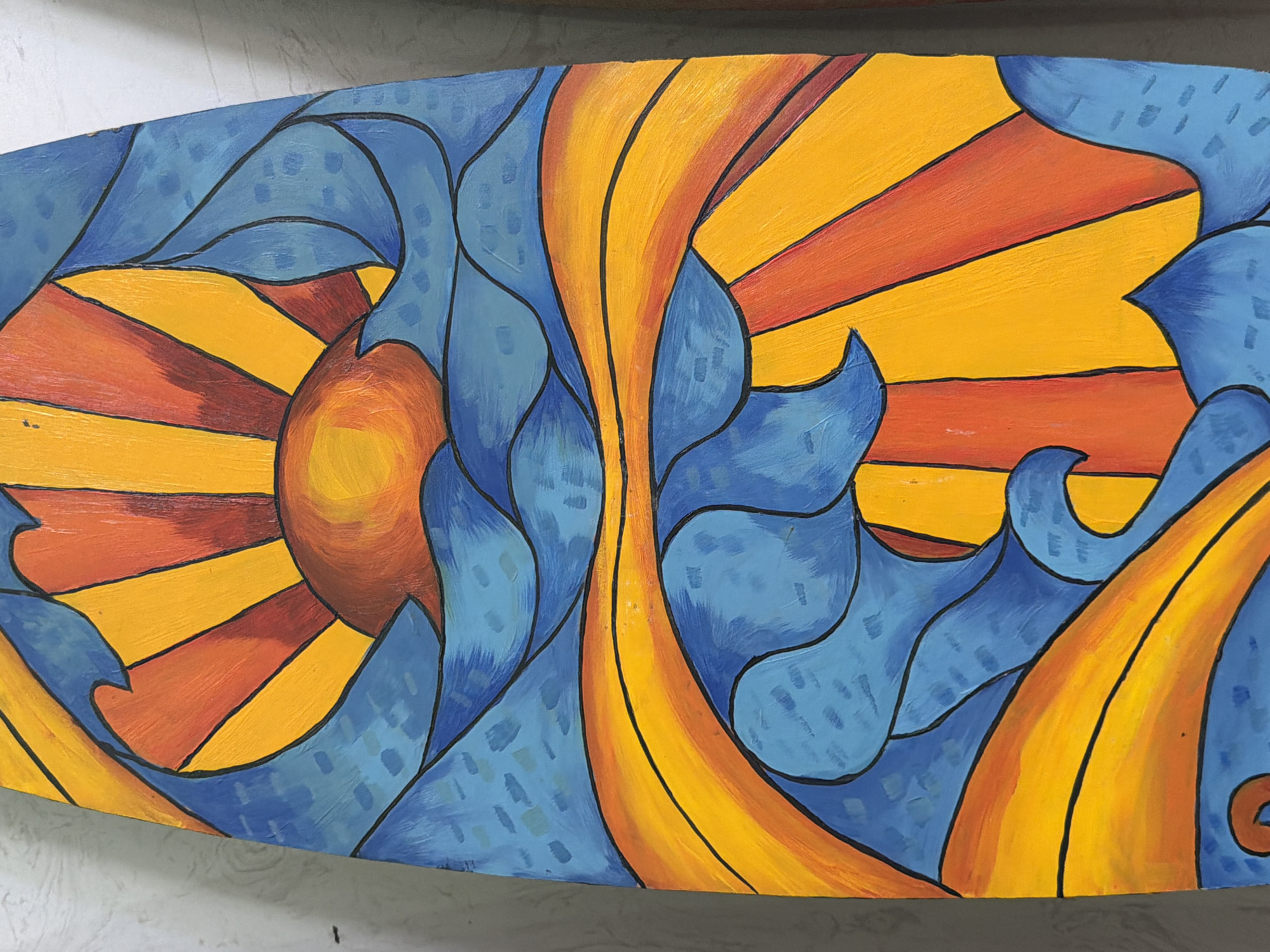 The image shows a close-up of a colorful, abstract painting on what appears to be a surfboard. The artwork features stylized waves in shades of blue, with a bright yellow and orange sun rising behind them. The sun's rays are depicted as bold, radiating lines of orange and yellow. The painting uses thick black outlines to define the shapes, giving it a stained-glass effect. The background is a textured, light-colored wall, suggesting the artwork is either displayed indoors or leaning against a wall. There are no people or animals visible in the image, and the specific location in Đà Nẵng, Vietnam is not discernible from the provided image.