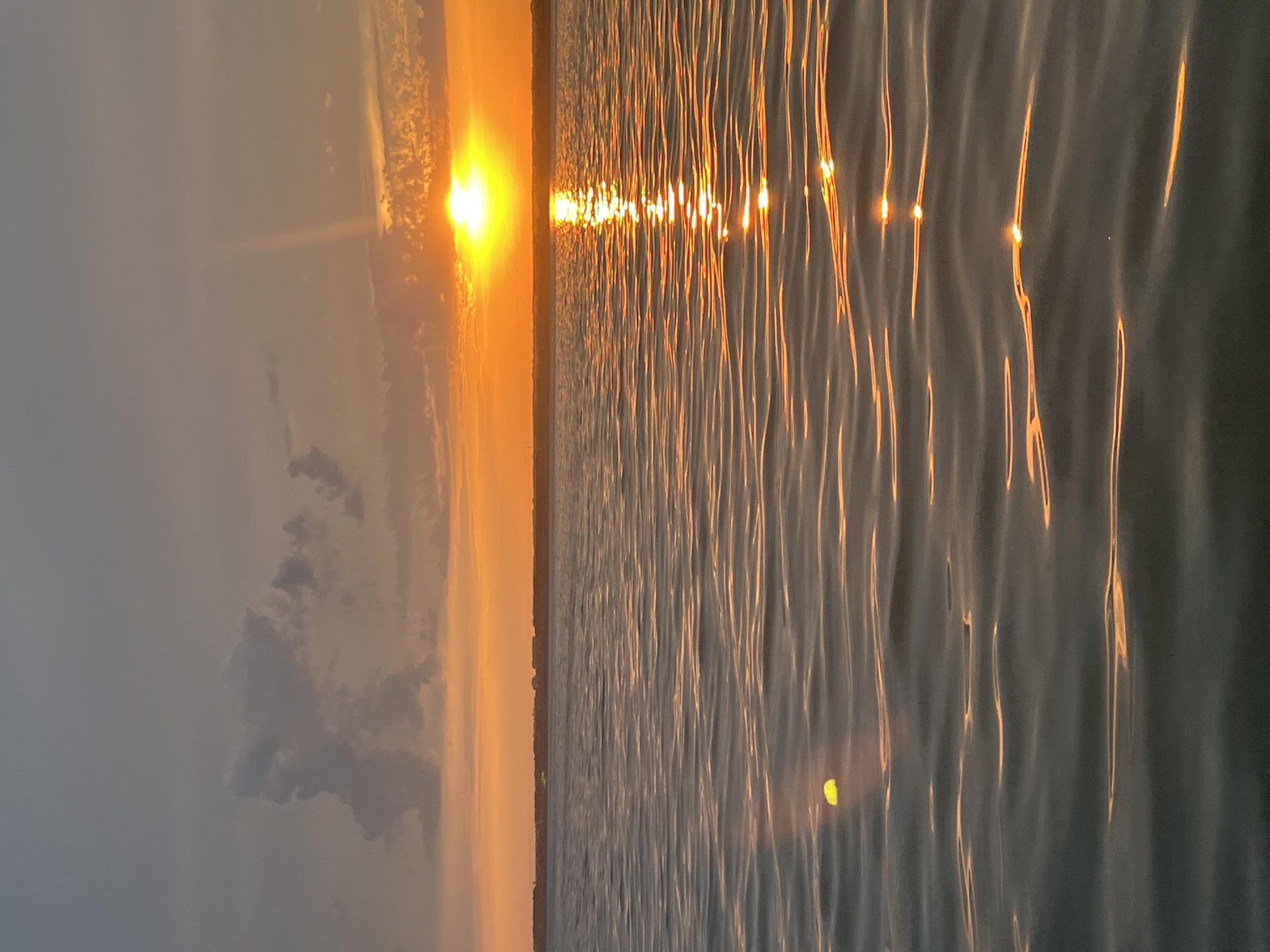 The image depicts a tranquil scene at sunset over a body of water, likely a lake or sea, in Beregovoe, Russia. The sky is a gradient of colors, with the sun, a brilliant orange orb, low on the horizon. Rays of sunlight pierce through scattered clouds, casting a warm glow across the scene. The water's surface is gently rippled, reflecting the vibrant hues of the sunset. In the distance, a faint silhouette of a shoreline with structures can be observed. A single, small, bright yellow object is visible floating on the water's surface in the foreground. The overall atmosphere is one of peacefulness and natural beauty.