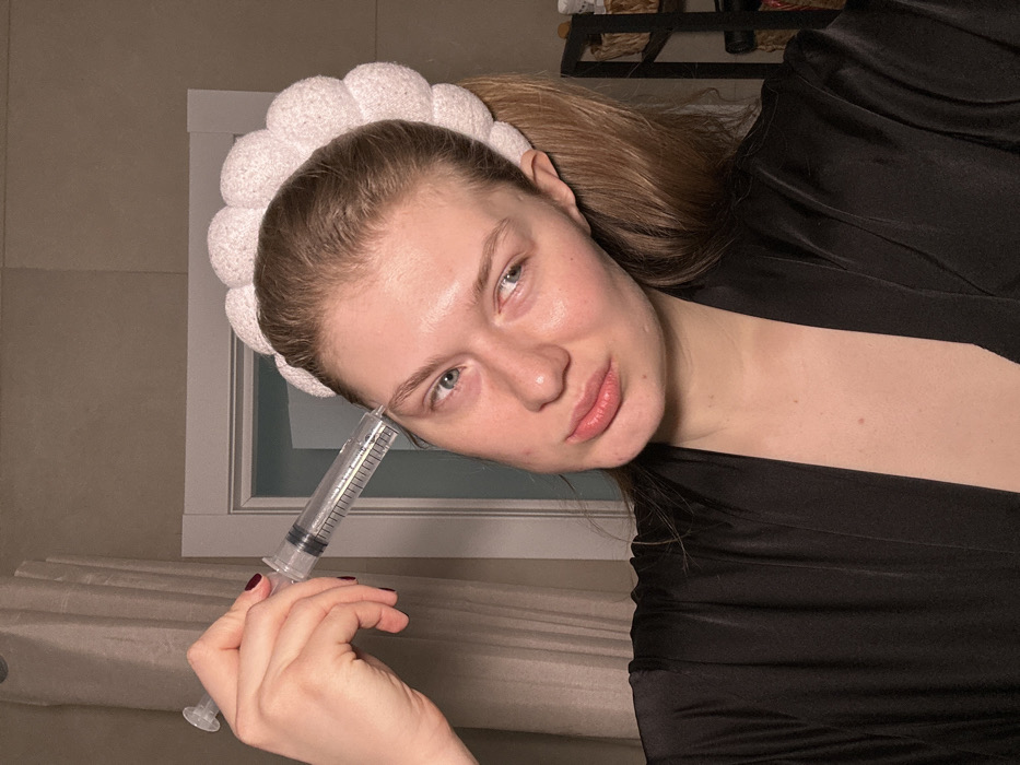A female subject, appearing to be an adult, is depicted indoors in Nikol'skii, Russia. She possesses light skin, light-colored eyes, and light brown hair gathered back and secured by a white, textured headband. She wears a dark-colored robe. Her right hand holds a large, clear plastic syringe, devoid of a needle, which contains a clear liquid and features visible measurement markings. The tip of the syringe is positioned against the skin above her left eyebrow. The subject's left eyebrow is raised, and her mouth is slightly pursed, directing a posed expression towards the viewer or camera. The background includes a white door frame, a light-colored curtain, and partially visible shelving containing woven baskets or containers. The environment is illuminated by artificial light.