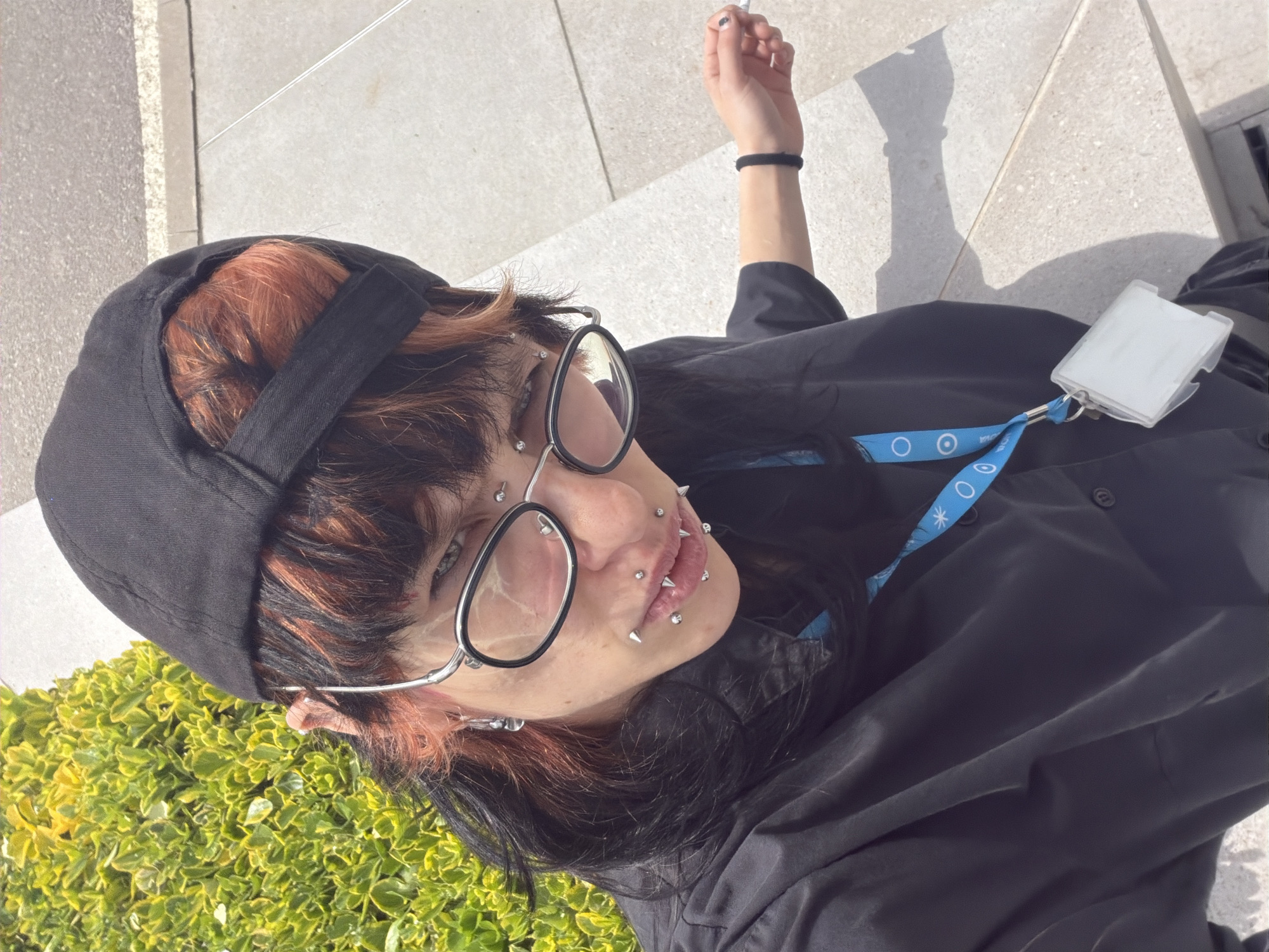 An overhead, high-angle shot captures a person outdoors in Busmantsi, Bulgaria, under bright, direct sunlight, evidenced by crisp shadows. The subject is a young adult with dark hair and prominent red/orange highlights, styled with bangs. They wear a black baseball cap backward and a black long-sleeved shirt.
Their face is adorned with several piercings, including studs on the cheeks and spiked labret piercings around the mouth. They wear silver-framed round glasses and look directly into the camera with a neutral expression. A black elastic hair tie is visible on their left wrist, and their nails are painted black. A blue lanyard, featuring white circular logos and an asterisk pattern, hangs from their neck, attached to a translucent white ID card holder that appears empty or contains an unreadable card.
The background shows light-colored paved ground, likely concrete or stone tiles, and a patch of vibrant green, low-lying bushes on the left side of the frame. The strong, clear lighting indicates it is daytime. No discernible text is visible on the lanyard or ID holder.