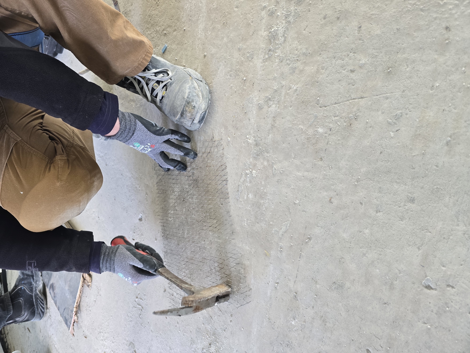 A person wearing work boots, brown pants, and a black top is shown kneeling on a concrete surface, securing wire mesh to the concrete with a hammer. The person is wearing gloves. The location is Unknown, Unknown.