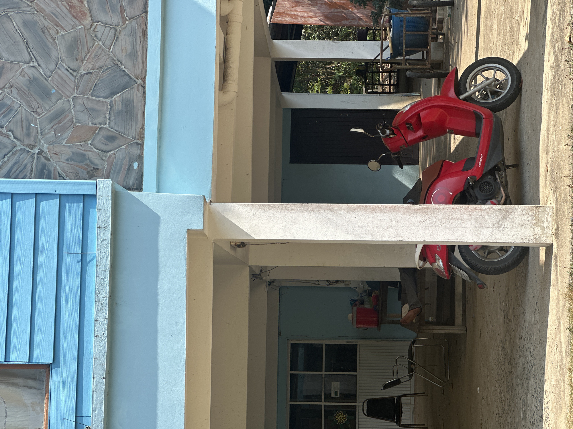 The image shows a red motorcycle parked in a covered outdoor area of a building in Ao Hin Khok, Thailand. The building has light blue walls and a textured stone facade. There are windows and doors visible in the background, along with chairs and other objects. The ground is concrete.