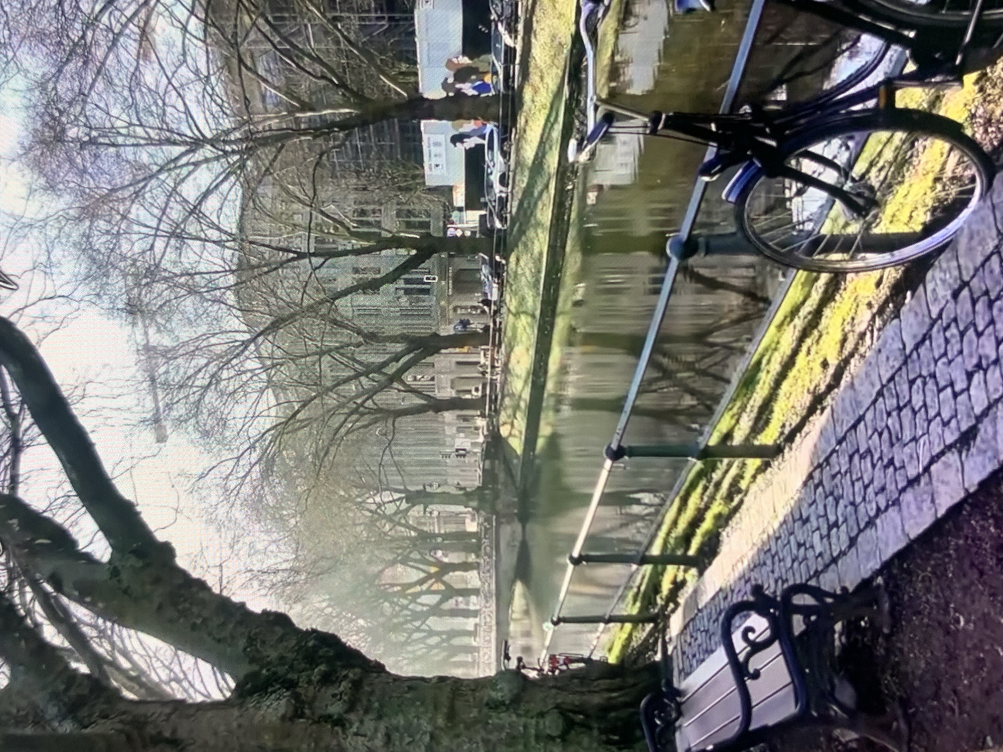 The image shows a canal lined with trees and buildings. A paved walkway runs alongside the canal, with a bench and a bicycle placed on it. A bridge crosses the canal in the background, and several cars are parked along the opposite bank. The sun is shining, casting long shadows on the walkway. The scene appears to be a peaceful urban setting.
The provided image and its crops do not contain any discernible information that would suggest the location is Stavropol, Russia, or any specific city. Therefore, I cannot fulfill the request to describe the location as Stavropol, Russia.