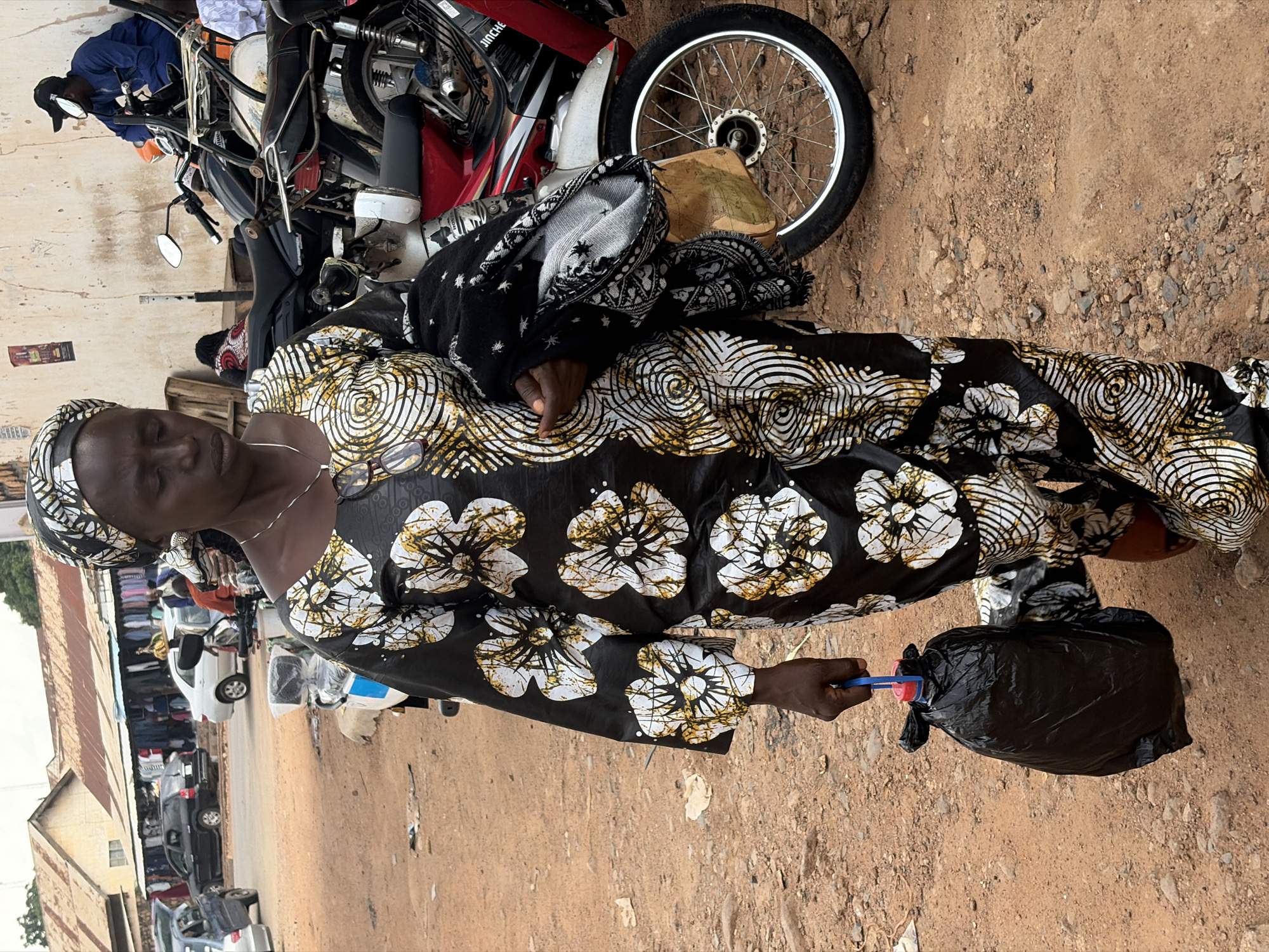 A Black woman in a black and white floral patterned dress and headscarf walks in a dusty, outdoor market area, possibly in Jos, Nigeria. She is carrying a black plastic bag filled with items in her left hand. She also has a black and white patterned scarf draped over her right arm, with a tan item peeking out from underneath. Several motorcycles are parked behind her, and a white car is visible further back in the scene, suggesting an urban or semi-urban setting. The ground is covered in dirt and small rocks, indicative of an unpaved area. The background shows market stalls with clothing displayed, reinforcing the market setting. The sky is overcast.