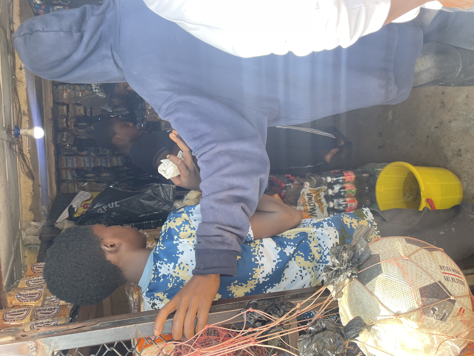In Barakin Lerawi, Nigeria, a group of individuals is observed at a roadside stall. In the foreground, a young boy wearing a blue and yellow camouflage t-shirt stands to the left, his right arm extended towards the viewer. He is holding a white object, possibly a piece of paper or cloth. To his right, a person in a blue hooded sweatshirt, facing away from the camera, also holds a similar white object in their right hand. Behind them, the stall is filled with shelves stocked with various packaged goods, including loaves of bread and snacks. A lit lightbulb hangs from the ceiling. A red mesh net containing a white and black soccer ball is suspended to the left of the stall. Further within the stall, another person is visible in the background, partially obscured. A yellow bucket filled with a brownish liquid sits on the ground to the right of the soccer ball. The ground appears to be concrete. The overall scene depicts a commercial transaction or interaction taking place at a local market setting.