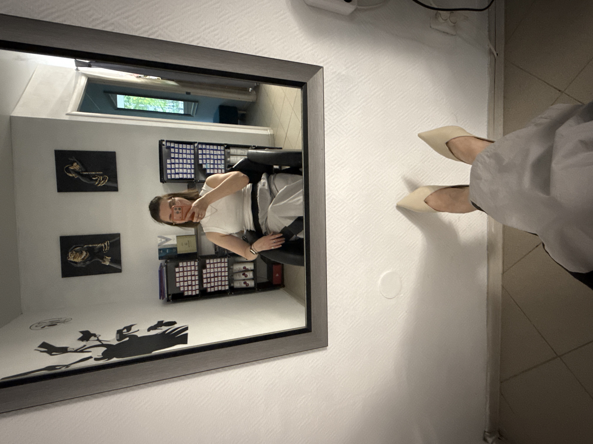 A woman in a white top and light-colored pants is sitting in a black chair, taking a photo of herself in a mirror. The room contains artwork on the walls and shelving units holding various items. The reflection in the mirror shows a window with greenery visible outside. The foreground shows the lower legs and shoes of a person standing near the mirror. The location of this scene is Chaikovsky, Russia.