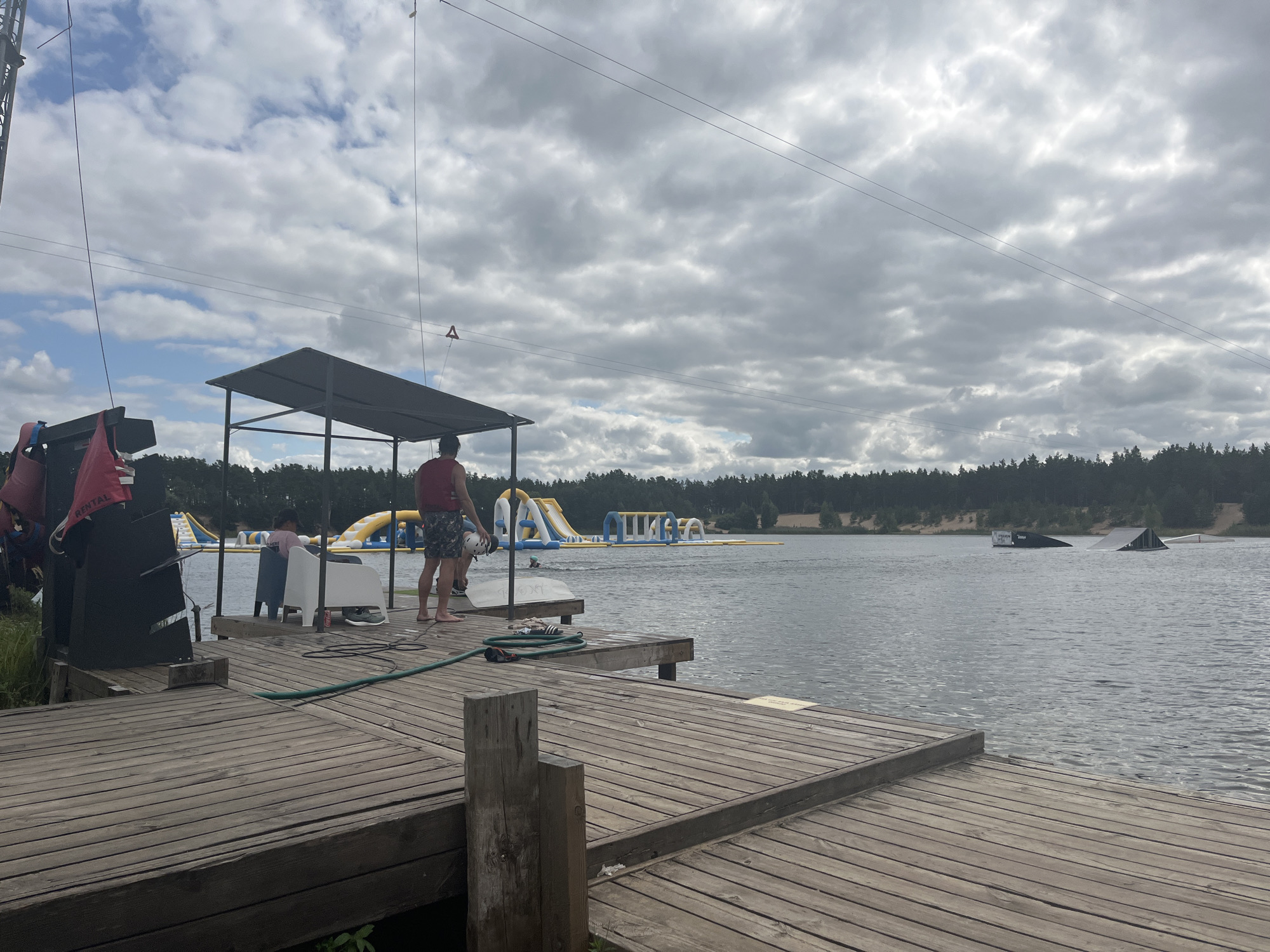 The image shows a wooden dock on a lake, with a cable wakeboarding park in the background. The park features inflatable obstacles and ramps. In the foreground, there are several people on the dock, some wearing life vests. One person is holding a helmet. The sky is cloudy. The location is Männiku, Estonia.