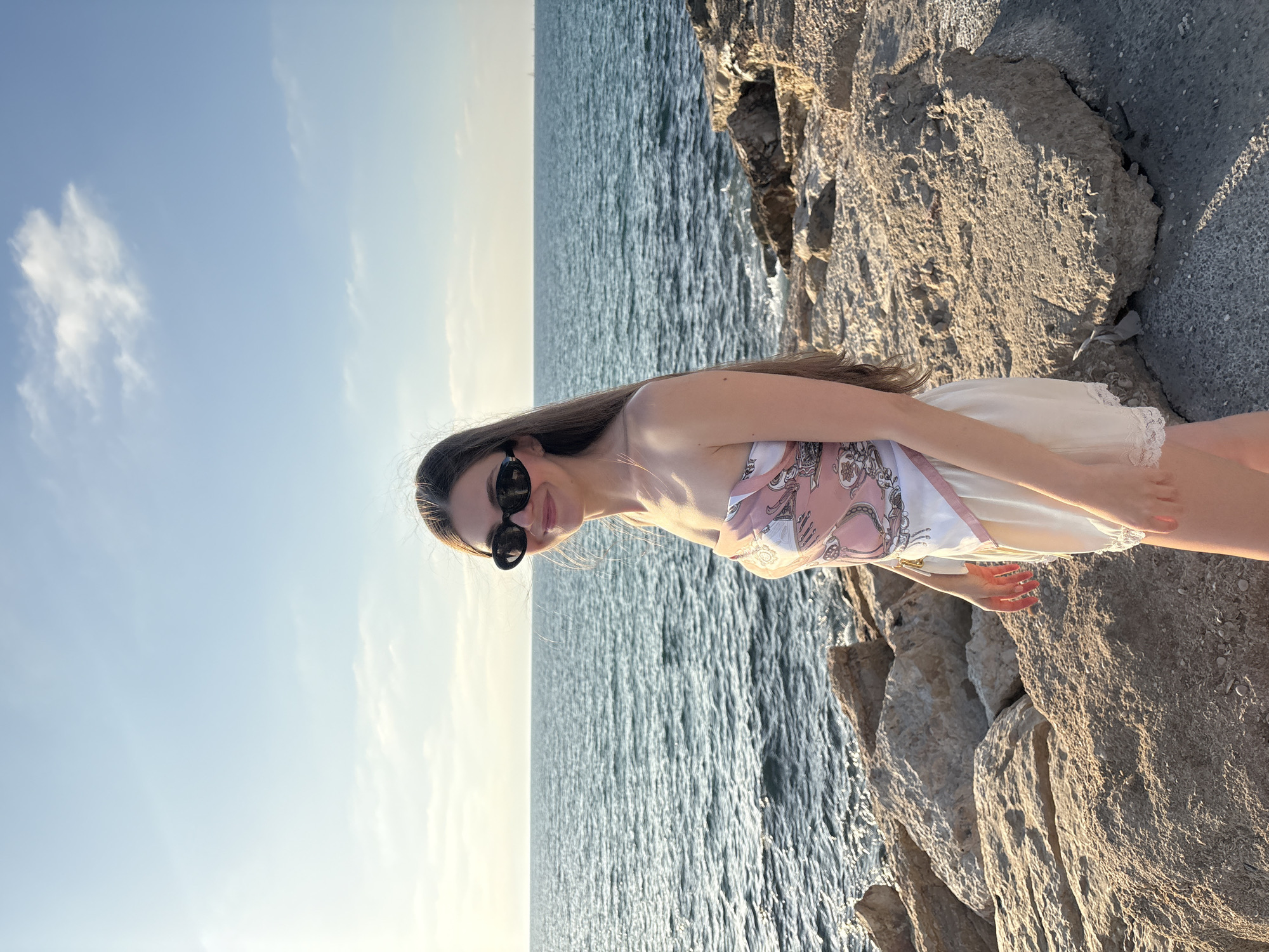 A young woman with long, brown hair and sunglasses stands on a rocky shore overlooking the Mediterranean Sea. She is wearing a strapless pink and white patterned top and a light-colored skirt. The sun is setting, casting a warm glow on the scene. The location is the Tel Aviv coastline, Israel, characterized by the rocky shoreline and the vast expanse of the sea under a clear sky.