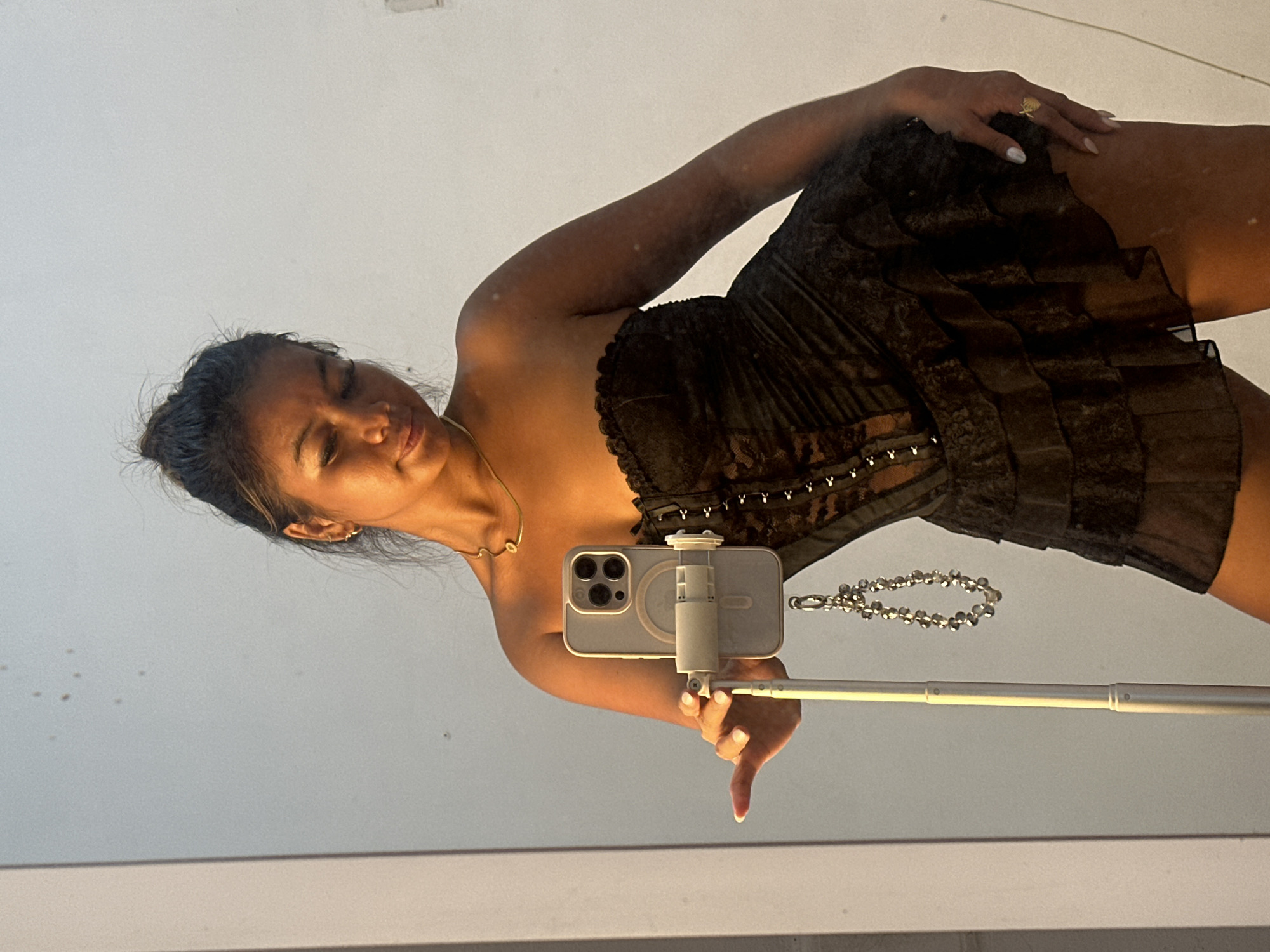 A woman is holding a smartphone attached to an extended selfie stick, likely taking a picture in a mirror. The woman is wearing a strapless black dress with a tiered, ruffled skirt. She is also wearing a gold necklace and a gold ring. Celuk, Indonesia is not discernible from the image.