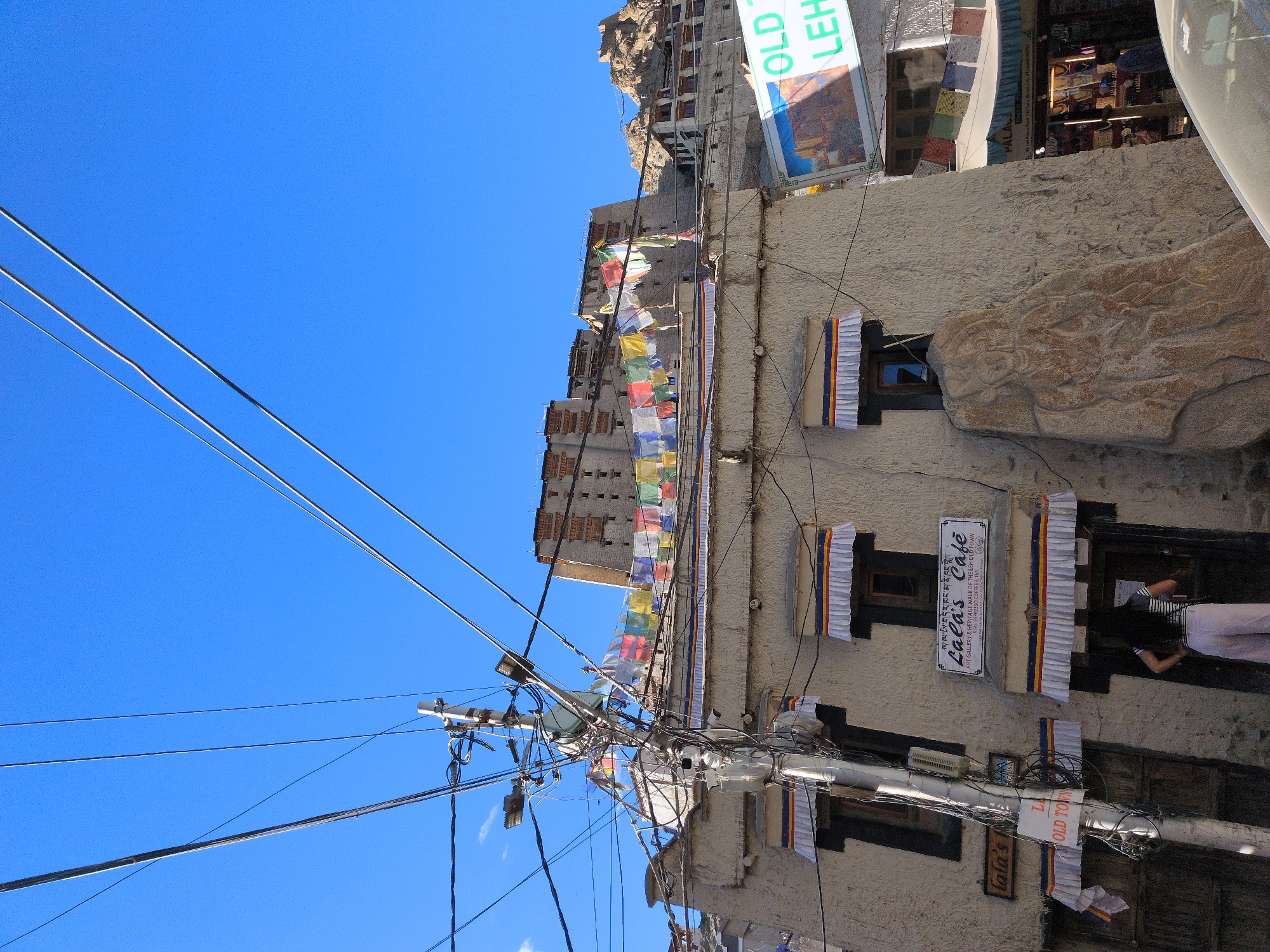 The image captures a street scene in Leh, India, featuring a stucco building with a sign that reads "Lala's Cafe" and "ART GALLERY & HERITAGE WALK OF THE LEH OLD TOWN." Prayer flags, strung across the street, flutter against a clear blue sky. Utility poles with numerous wires crisscross the frame, adding a sense of urban density. In the background, the historical Leh Palace and surrounding structures are visible, perched on a hillside. A person wearing a striped top and light-colored pants stands at the entrance of the building, with their back to the camera. To the right, a shop with an awning displaying "PALACE" in large letters is visible, with goods displayed inside and a person partially obscured by the entrance. A large carved rock sits beside the cafe, adding an artistic element to the scene. The overall impression is one of a bustling, culturally rich, and somewhat eclectic urban environment.