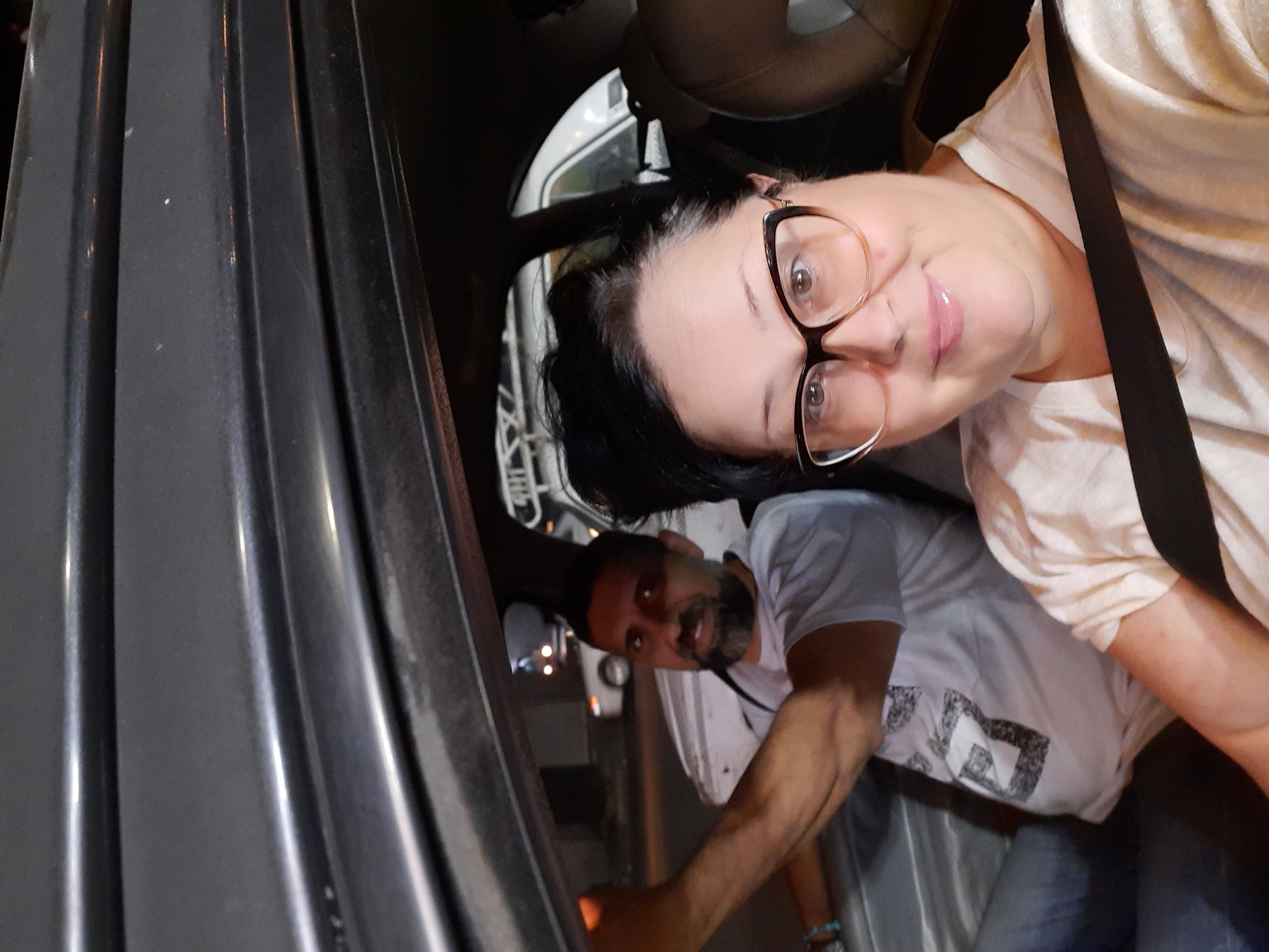 Two people are inside a vehicle. The person in the foreground is wearing glasses and a seatbelt. The person in the background appears to be exiting the vehicle. The background shows a street with other vehicles and lights, suggesting a nighttime setting. No information suggests the location is Cairo, Egypt.