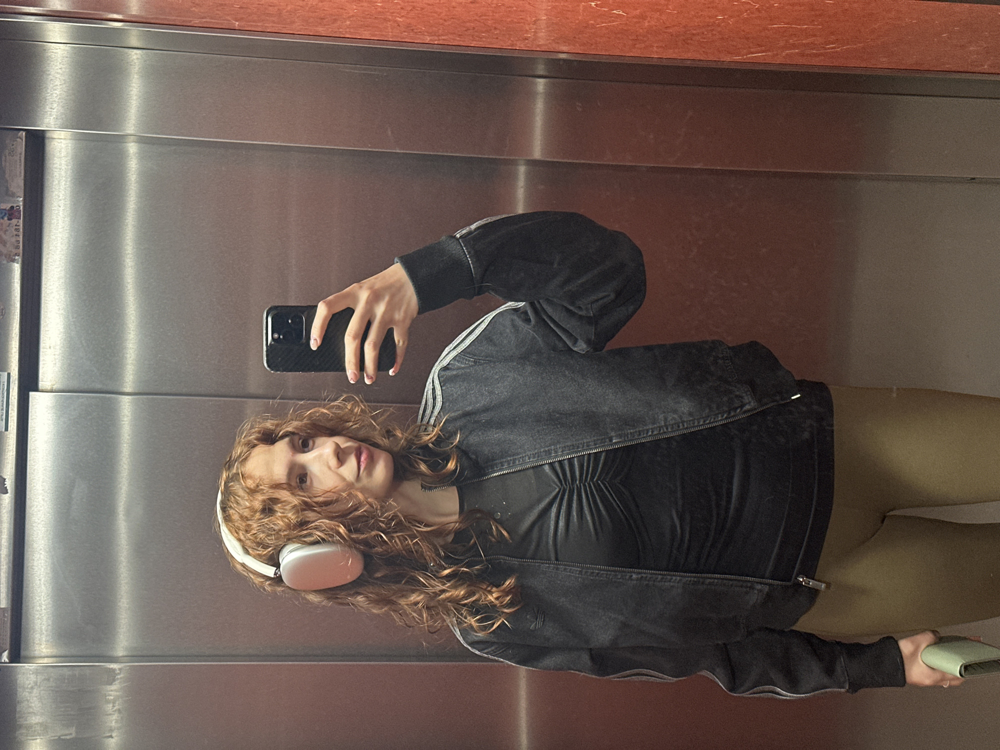A young woman with curly red hair is taking a selfie in an elevator. She is wearing white headphones, a black top, a black jacket with white stripes, and green leggings. She holds a black smartphone in her right hand and a green wallet in her left. The elevator has stainless steel walls. The top panel of the elevator is made from reddish-brown marble. There is no indication this image was taken in Novi Sad, Serbia.