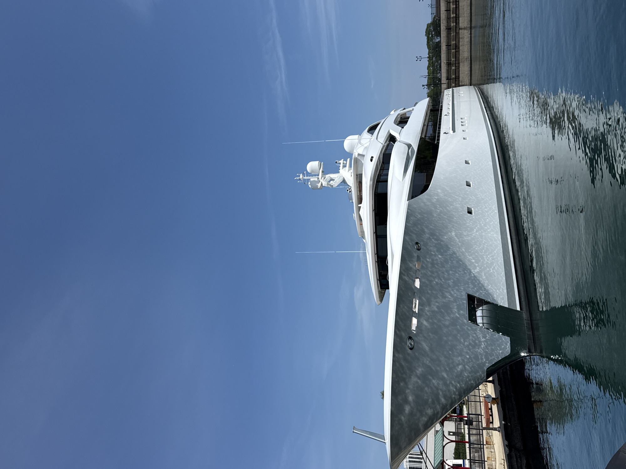 This media file shows a large, modern white yacht docked in a harbor. The yacht is the primary subject, with its sleek lines and multi-deck structure prominently displayed. The scene is set against a clear, bright blue sky with a few wispy clouds. The water is calm and reflects the yacht and the sky. In the background, a concrete seawall with some greenery and structures is visible, suggesting an urban or developed waterfront. The lighting indicates it is daytime, likely midday, with bright sunlight casting reflections on the yacht's hull and the water. The image is taken from a low angle, emphasizing the scale of the yacht. There is no visible text in the media. The setting appears to be a marina or a harbor, with the calm water and docked vessel indicating a peaceful scene.