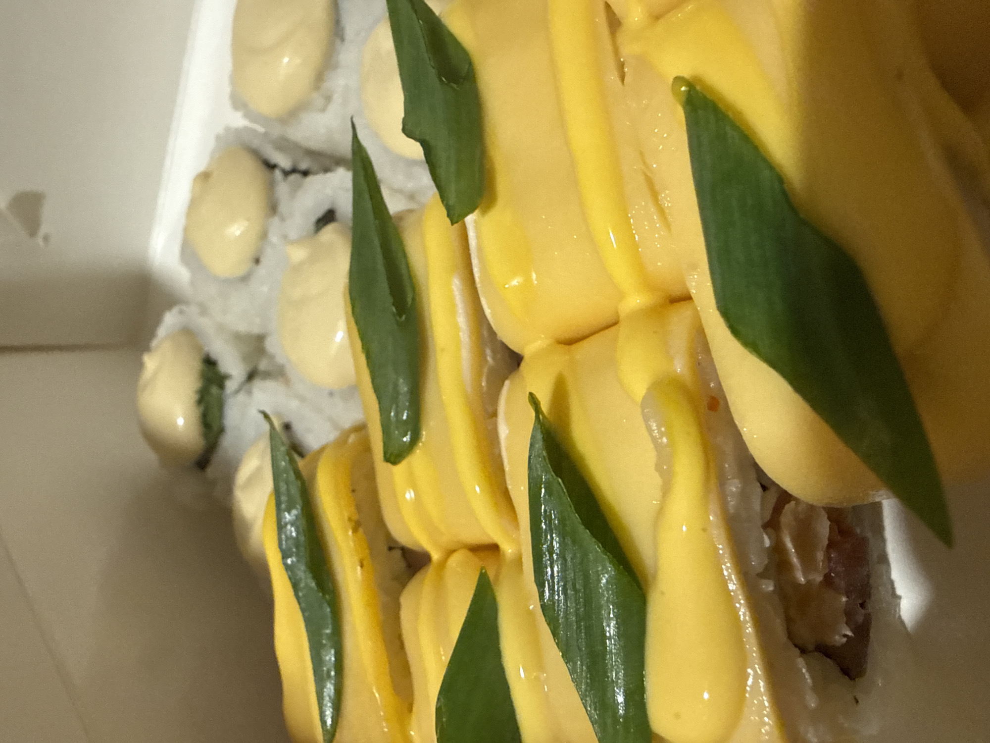 The image shows a close-up of sushi, likely in a takeout container. There are two distinct types of sushi. One type has white rice on the outside with dollops of a creamy, light-yellow sauce on top. The other type is topped with slices of a bright yellow fruit, likely mango, with similar creamy sauce drizzled over. Green leafy garnishes are placed on top of the mango sushi. The interior contents of the sushi are partially visible, appearing to contain some meat or fish. The image is taken from a slightly angled, close-up perspective. No people, locations, nor environment pertaining to the city of Dmitrov, Russia is seen in the provided image.