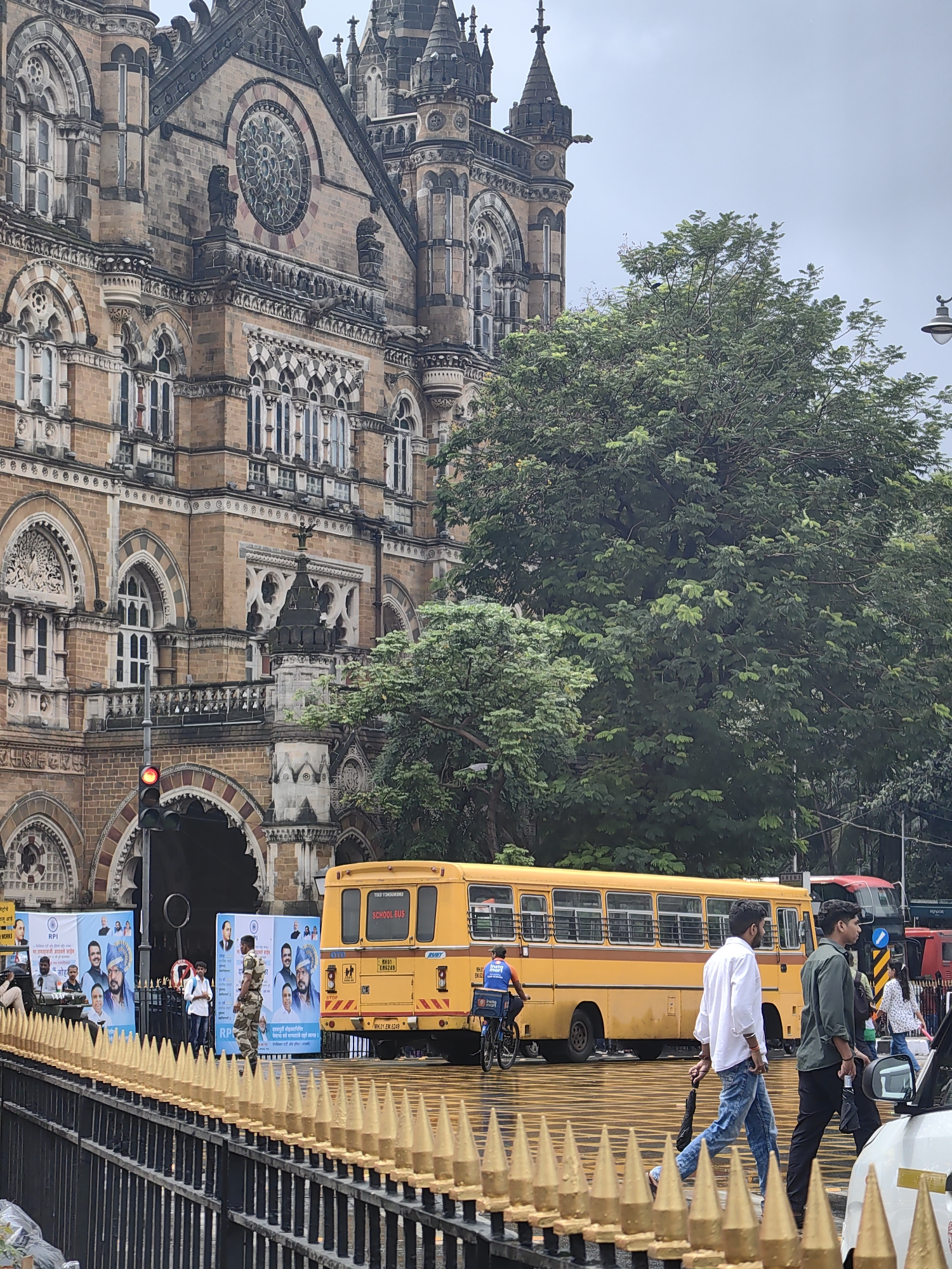 The image shows the Chhatrapati Shivaji Maharaj Terminus, a historic railway station in Mumbai, India. In the foreground, a black metal fence with golden pointed spikes lines the street. Behind the fence, several people are walking across a wet road, possibly from rain. A yellow school bus is prominently visible in the middle ground, along with other vehicles and people in the background. The iconic architecture of the railway station is visible in the upper portion of the image, with its intricate details and grand facade. The sky is overcast, suggesting cloudy or rainy weather.