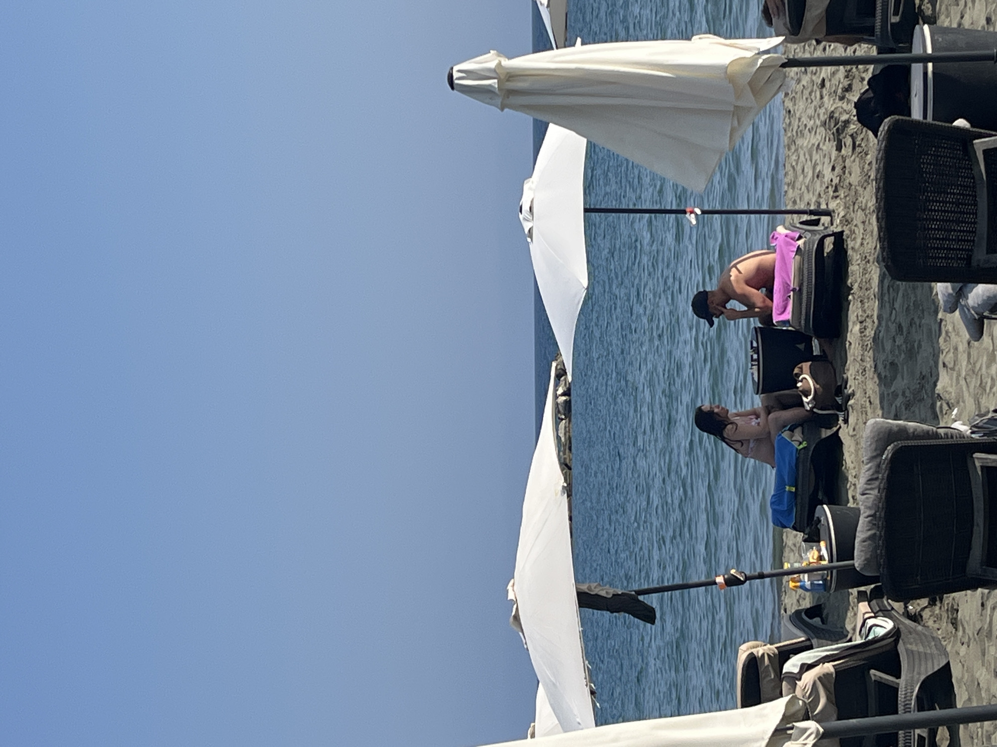 The image captures a daytime scene on a beach in Agios Tychona Community, Cyprus. Two individuals, a man and a woman, are seated on lounge chairs under separate umbrellas. They appear to be engaged in a conversation or activity at a small table placed between them. The woman is wearing a bikini, and the man is wearing swim trunks and a baseball cap. In the foreground, additional lounge chairs and umbrellas are visible, some of which are unoccupied. The sandy beach slopes down to the calm, blue Mediterranean Sea, with a clear sky overhead. The overall atmosphere suggests a relaxed beach setting.