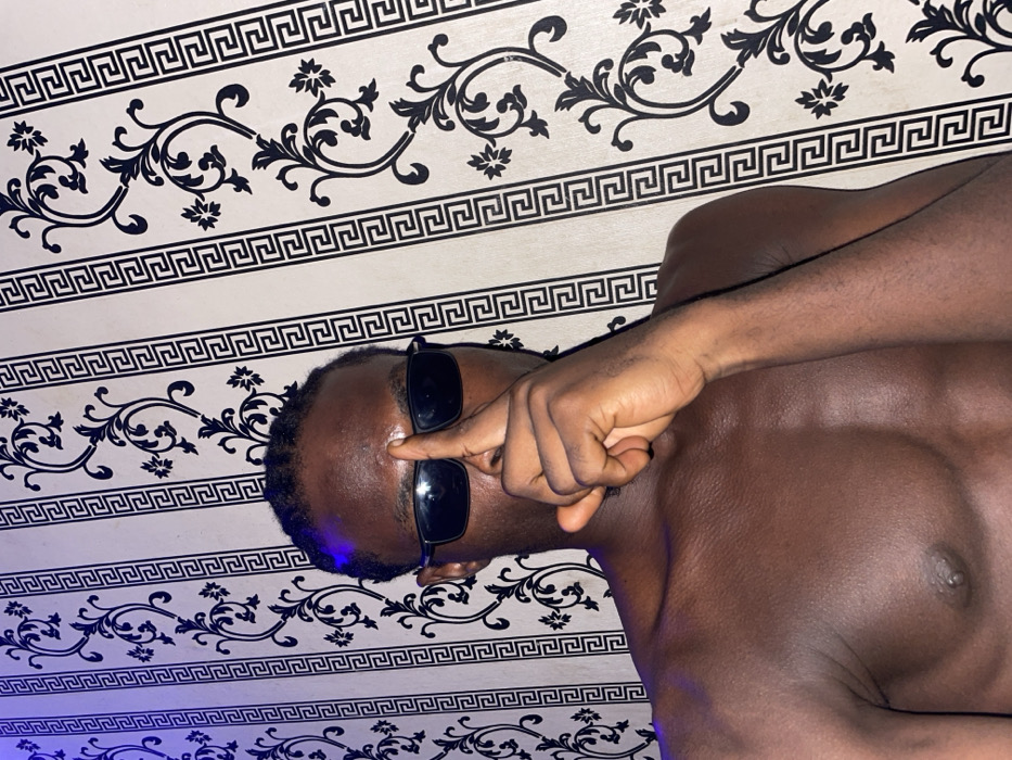 A shirtless, dark-skinned man with black sunglasses is lying down against a wall with ornate black and white wallpaper. He is holding his index finger up to his forehead as if making a point or emphasizing something. The lighting is dim, with a blue hue on the left side of the frame.