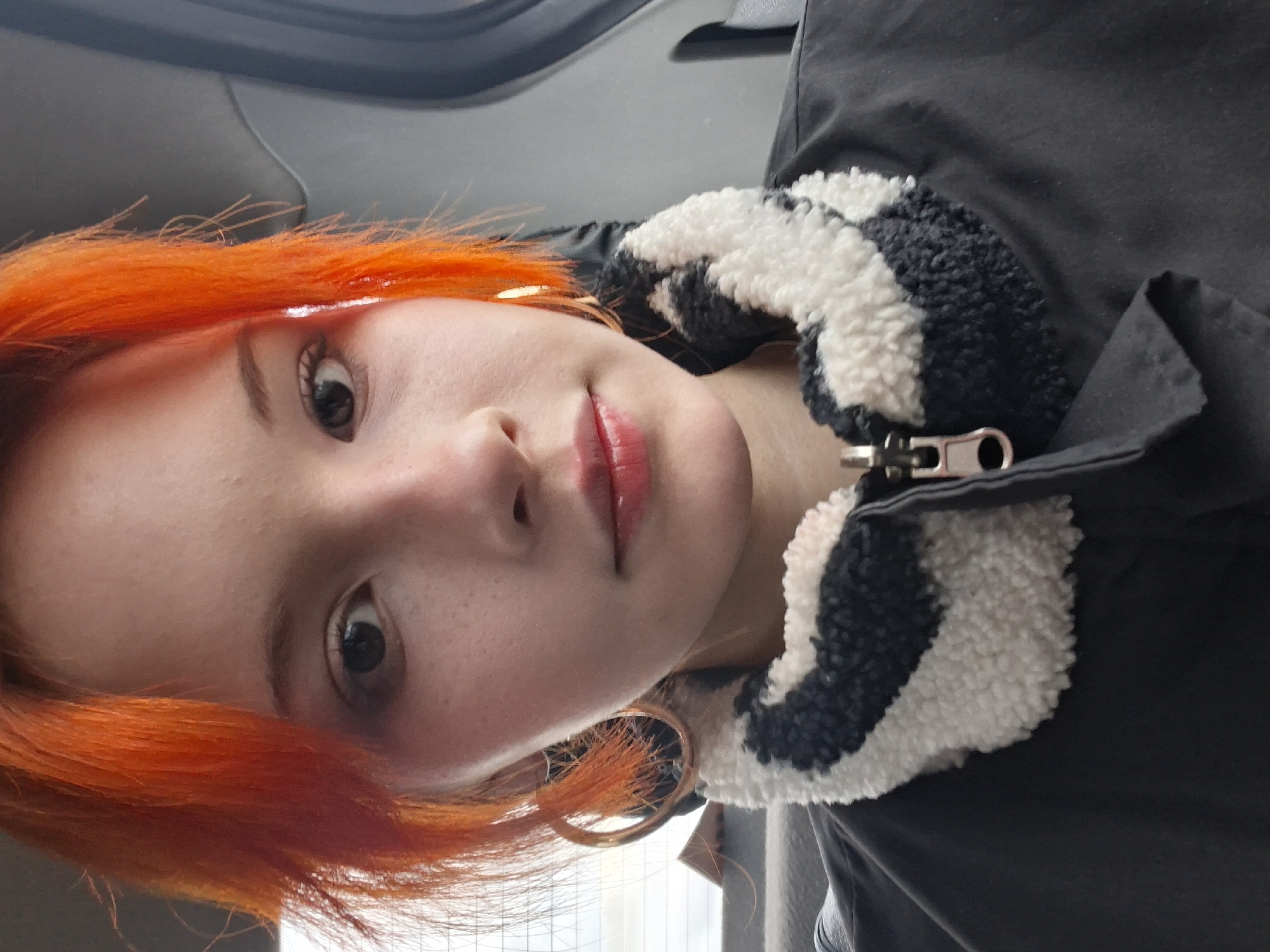 A close-up, rotated portrait shows a young woman with orange hair, large gold hoop earrings, and red lipstick. She is wearing a dark jacket with a black and white sherpa-lined collar. The background appears to be the interior of a vehicle. There is no visual information in the photo to confirm the location is Chaikovsky, Russia.