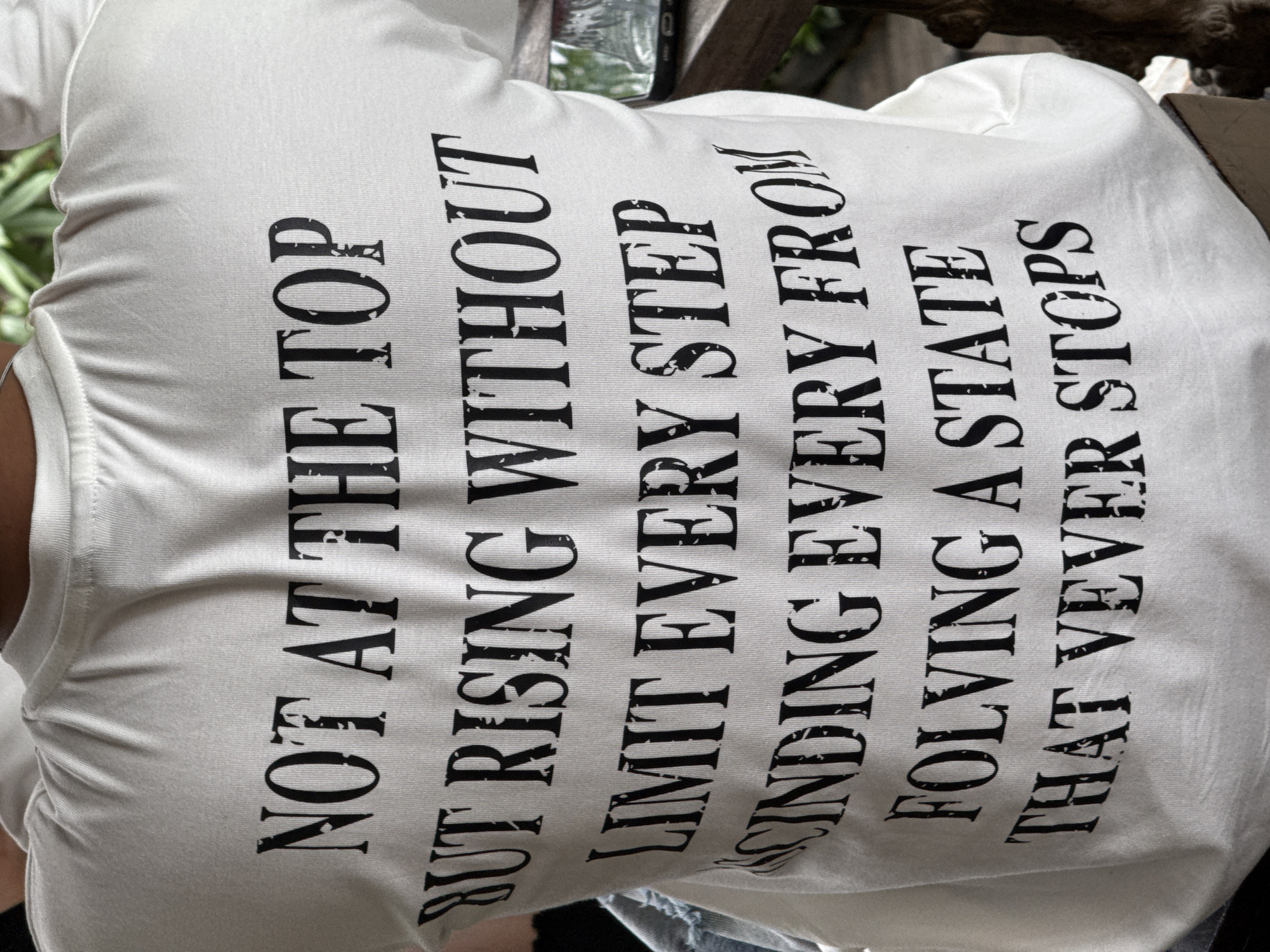The image shows a person wearing a white t-shirt with black text printed on the back. The text is in all capital letters and appears to be a quote. The t-shirt is slightly wrinkled. The person is partially visible from the back, with their head and shoulders out of frame at the top. The background is blurred and suggests an outdoor setting with foliage and a wooden surface. The scene is likely captured in Da Lat, Vietnam, as the text on the t-shirt seems to be a quote from a Vietnamese proverb. The overall lighting suggests it is daytime.