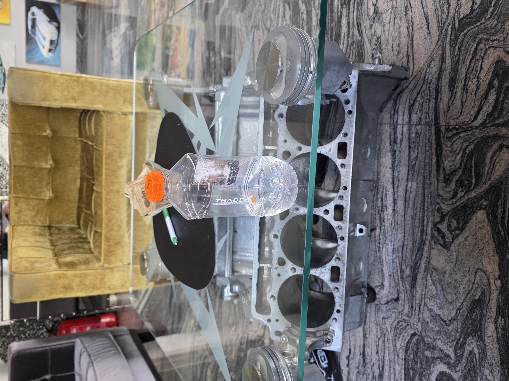 The image depicts an indoor scene, likely a commercial or exhibition space, featuring a unique glass-top coffee table. The table's base is constructed from an automotive engine block, with pistons visible within some of the cylinders. The glass surface of the table is partially obscured by a clear plastic bottle of liquid with an orange cap, a white marker with a green tip, and a small packet of sugar or creamer. In the background, a plush, gold-colored armchair is prominent, and to its left, a dark grey armchair is partially visible. Beyond the seating, a red fire extinguisher is mounted on a wall. The wall itself is adorned with artwork, including a painting of a classic white car with blue racing stripes. The floor is a polished granite with a distinctive black and grey marbled pattern. The overall impression is of a stylish, automotive-themed interior. The location is specified as Kladnitsa, Bulgaria.