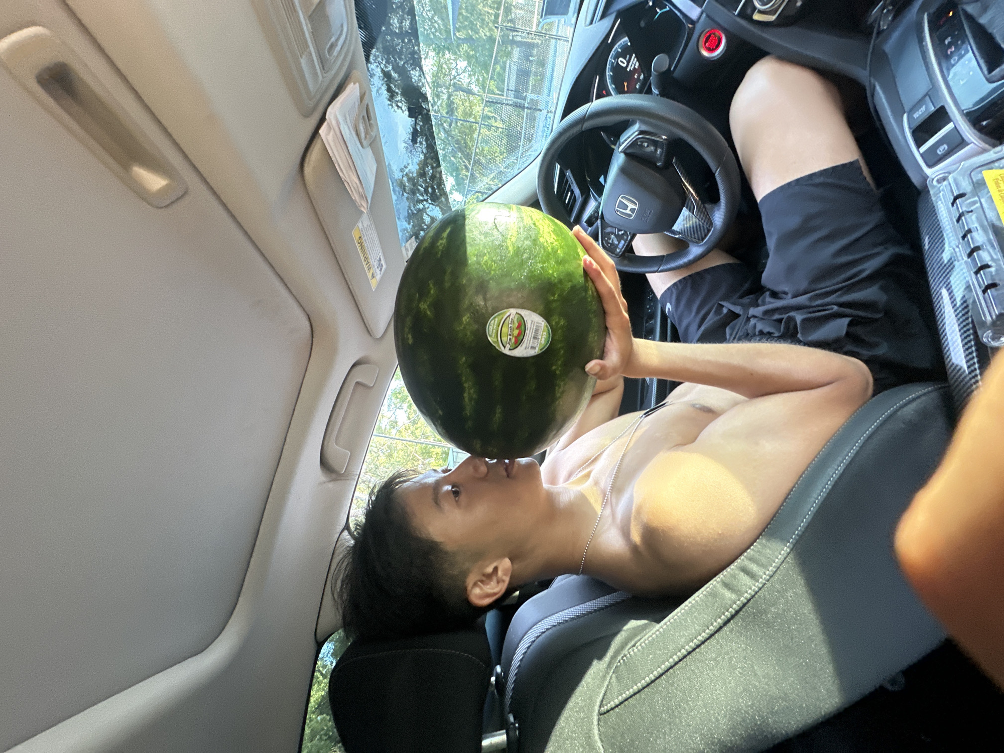 A person is lying in the passenger seat of a vehicle. They are shirtless and wearing black shorts. They are holding a large watermelon up to their face. The vehicle is likely parked somewhere in the city of University, United States of America, as the trees in the background are deciduous trees typical of that climate.